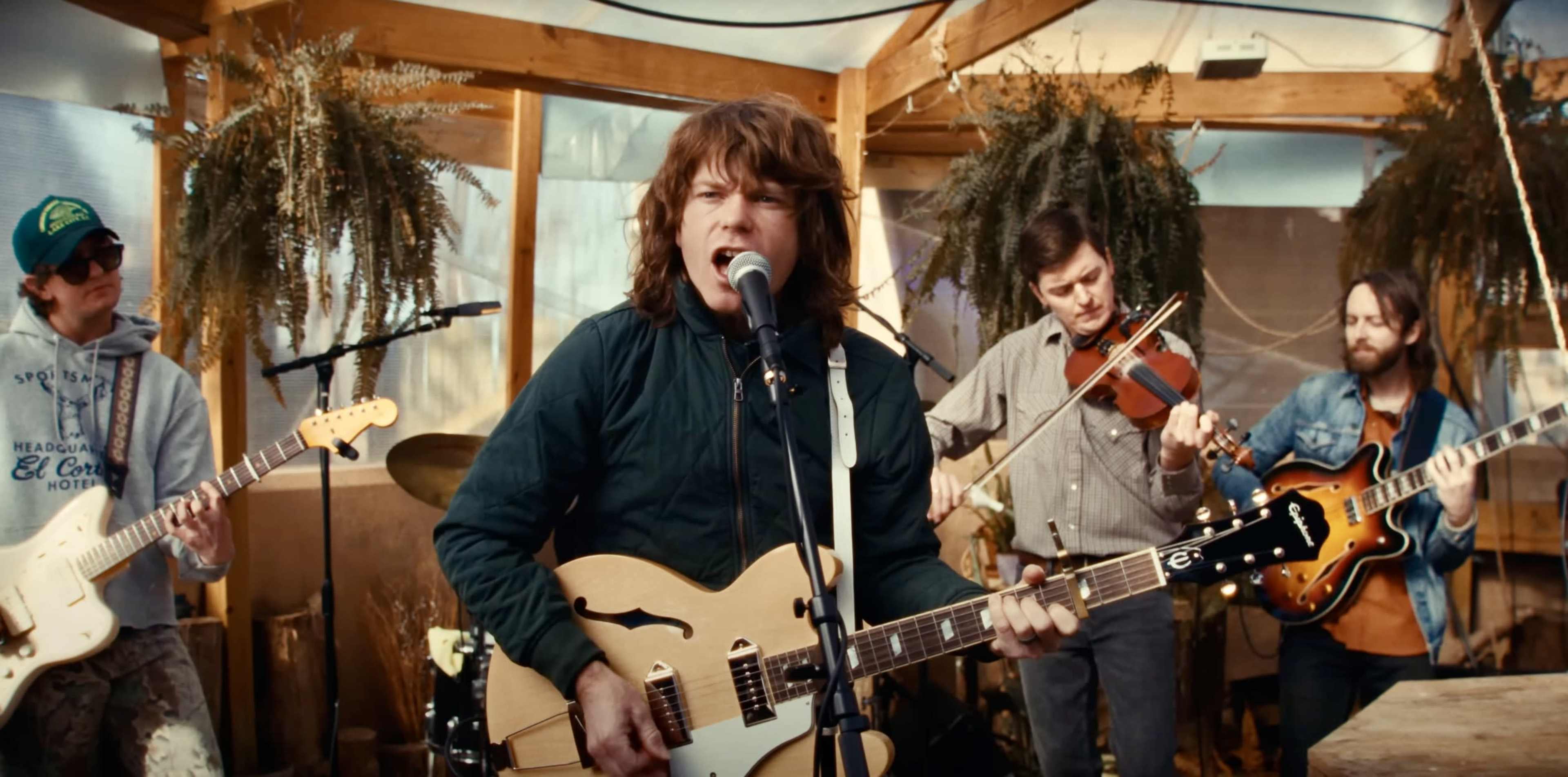 A band performs inside a greenhouse with instruments, including guitars and a violin, amid hanging plants.