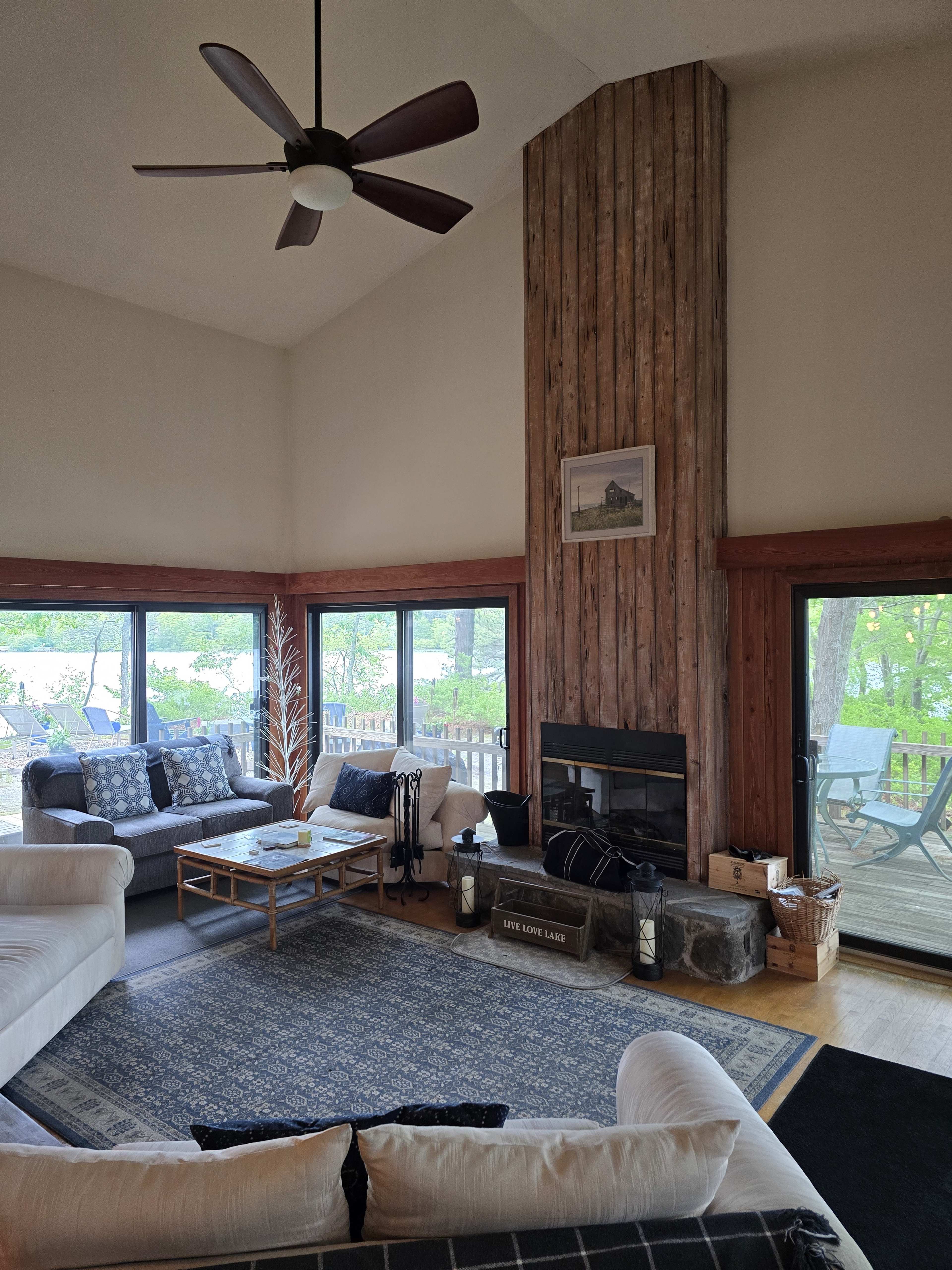 The image shows a spacious living room with large windows overlooking a deck and trees, featuring a tall wooden fireplace, a coffee table, and various seating arrangements.
