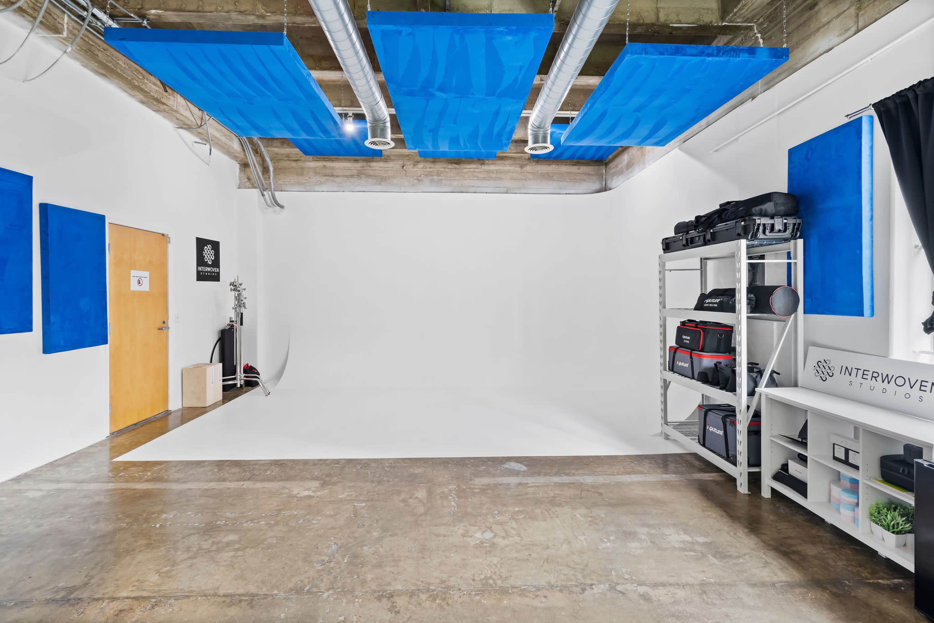 A bright photography studio with a white backdrop, a wooden door, blue soundproof panels on the ceiling, and shelves filled with camera equipment.