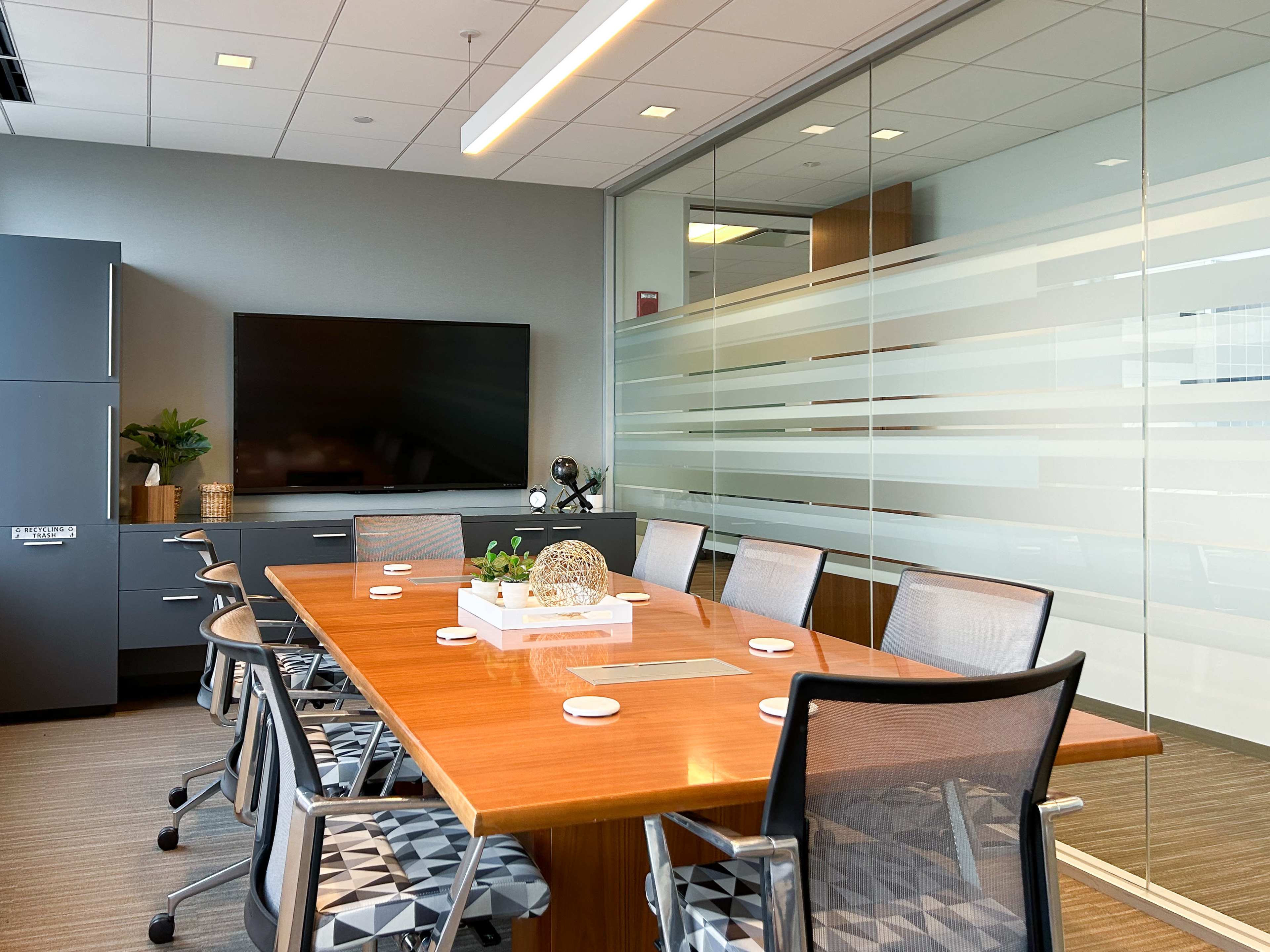 A modern conference room features a large wooden table surrounded by ergonomic chairs, with a television mounted on the wall and glass partitions.