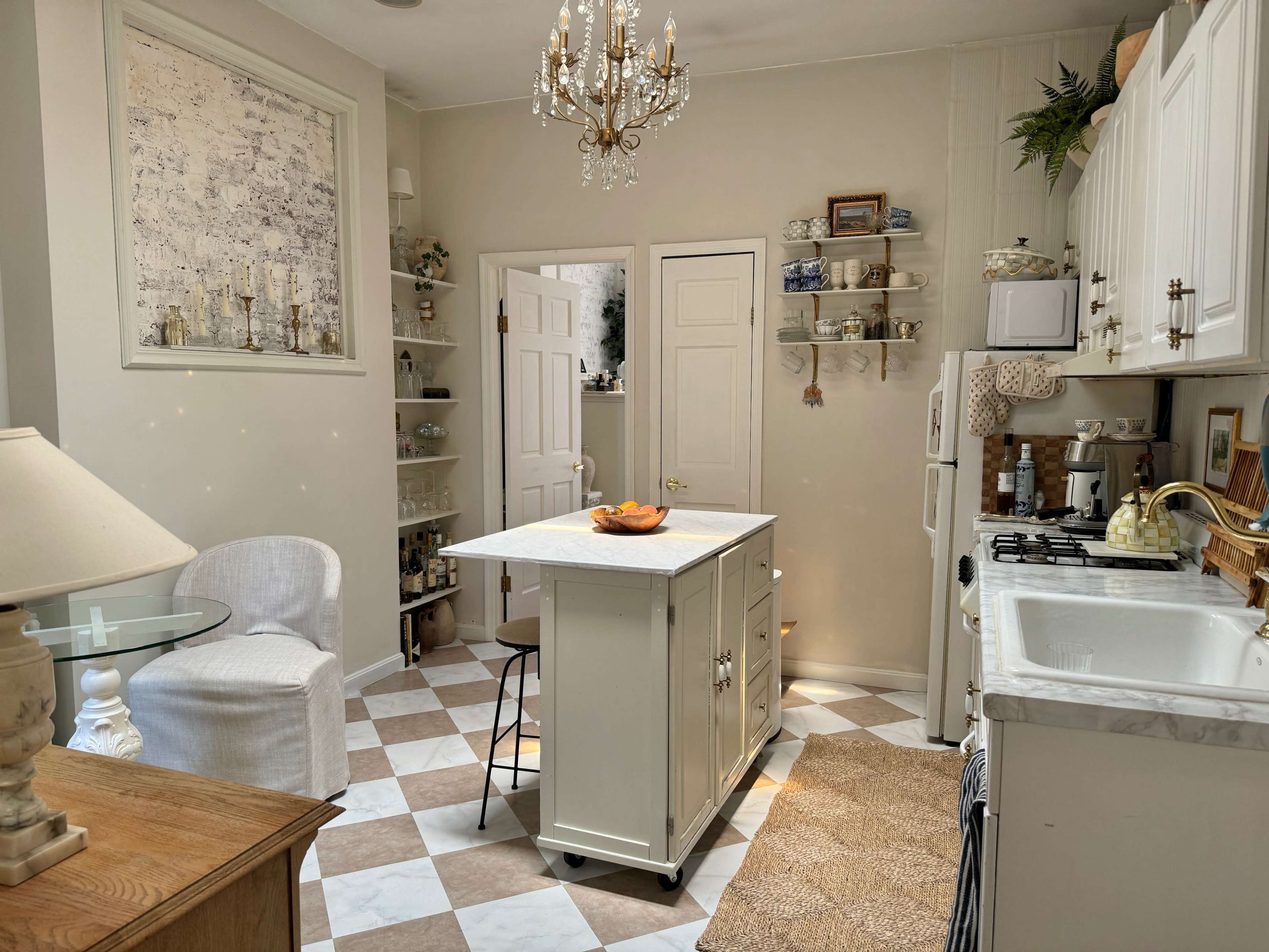 A well-organized kitchen features white cabinets, a central island with a bowl of fruit, and checkered flooring with shelves displaying decorative items.