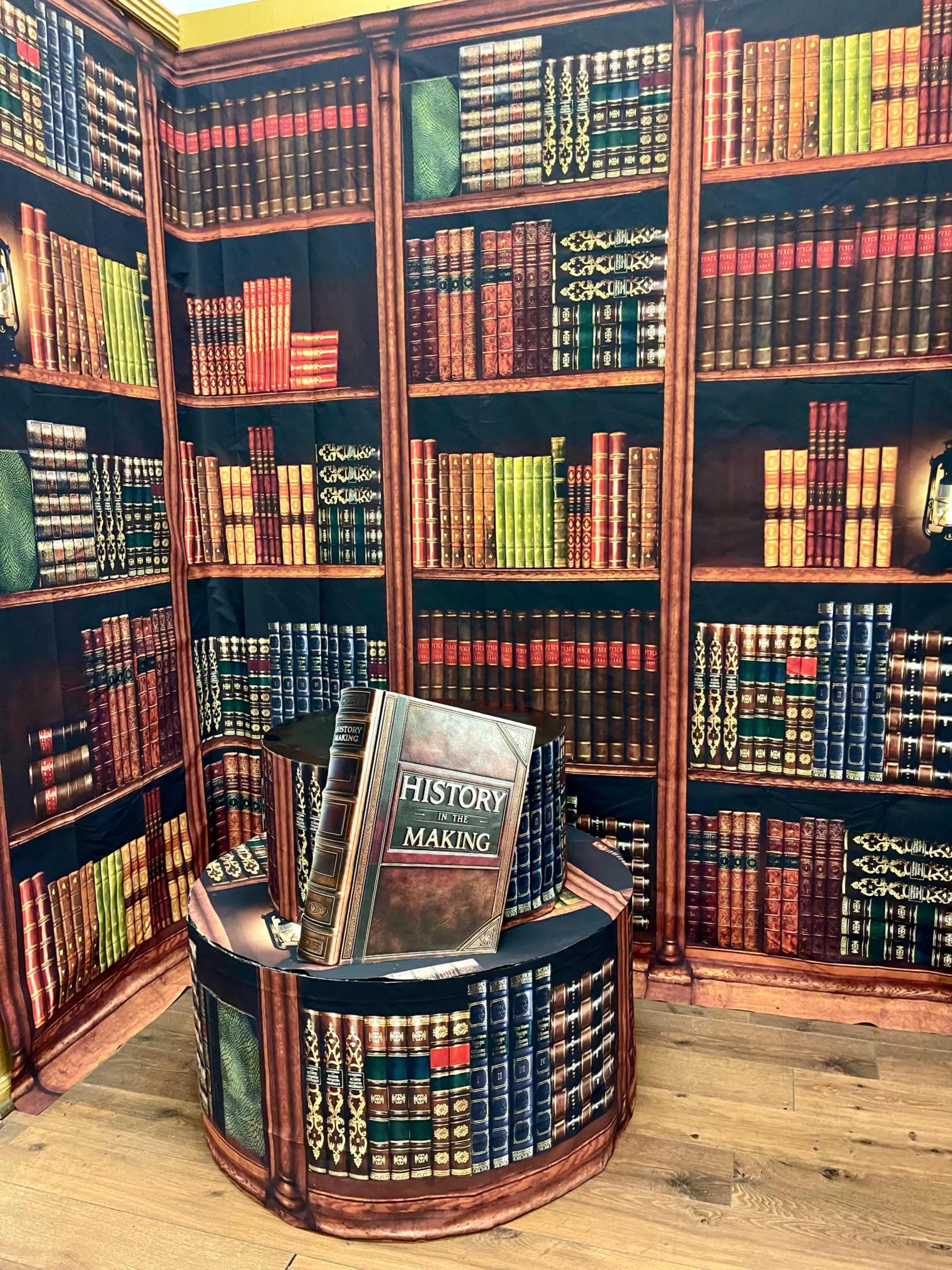 The image shows a decorated corner featuring a large bookshelf backdrop with artificially created books and a circular stand displaying a book titled "History in the Making."