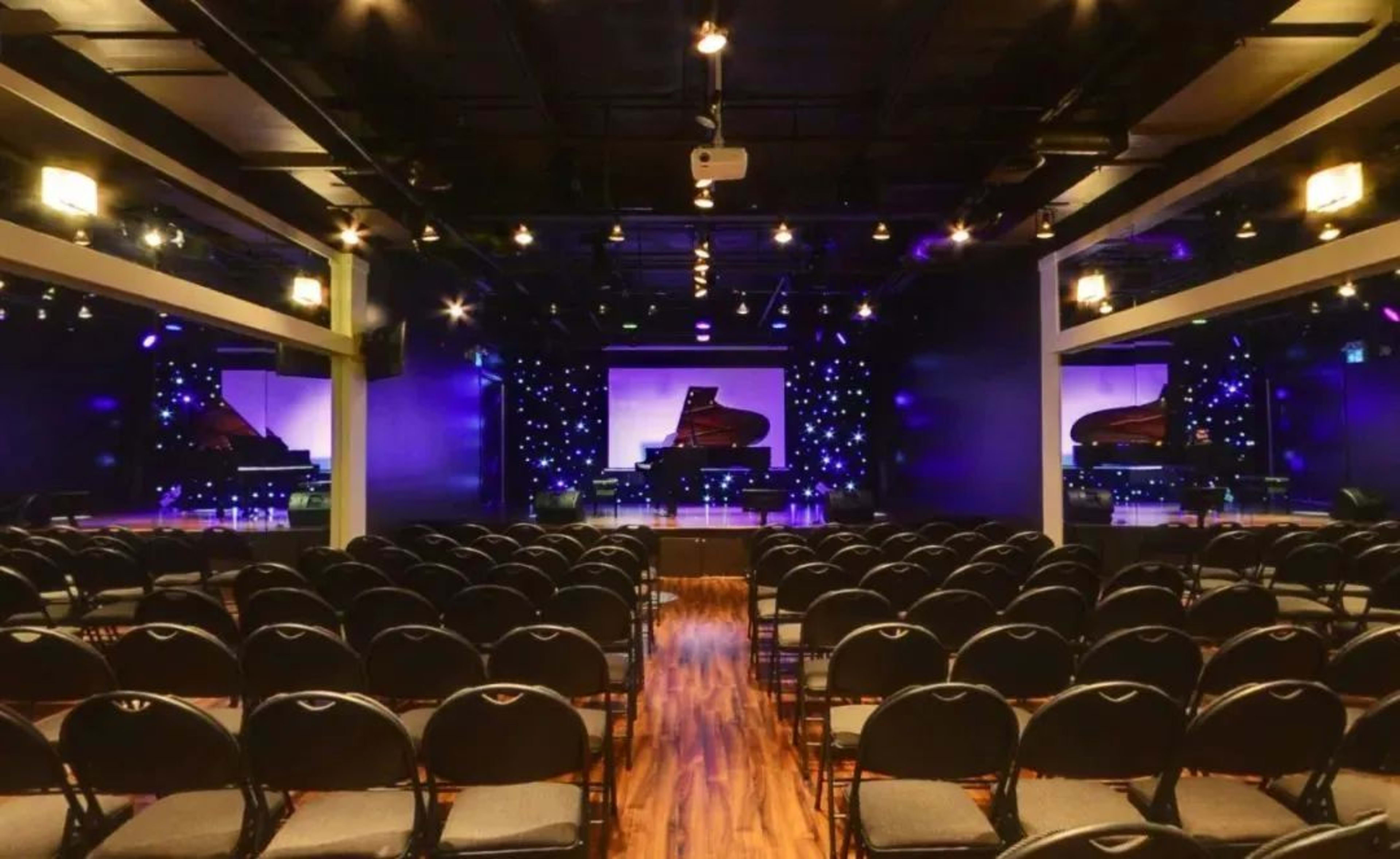 A concert hall is set up with rows of empty chairs facing a stage featuring pianos and a backdrop illuminated by purple lights.