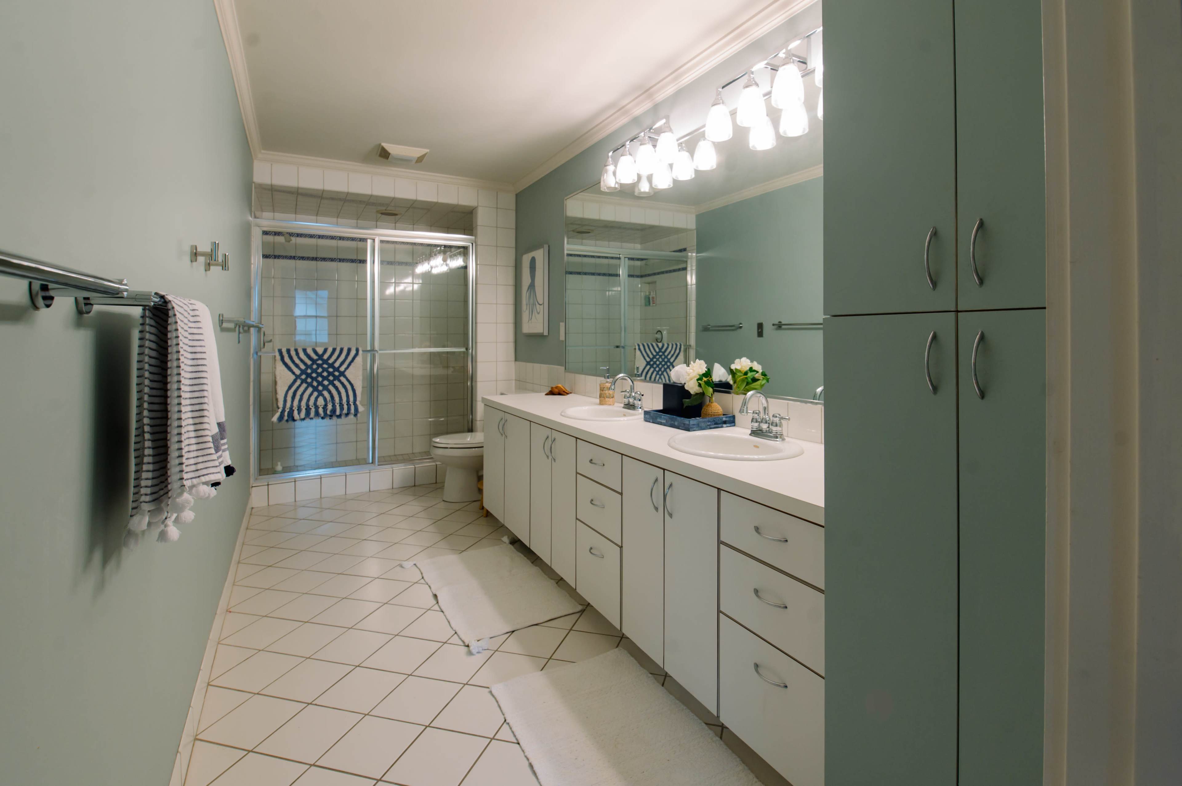 The image shows a spacious bathroom with mint green walls, white cabinetry, and a tiled floor, featuring a shower area and a large mirror above the sink.