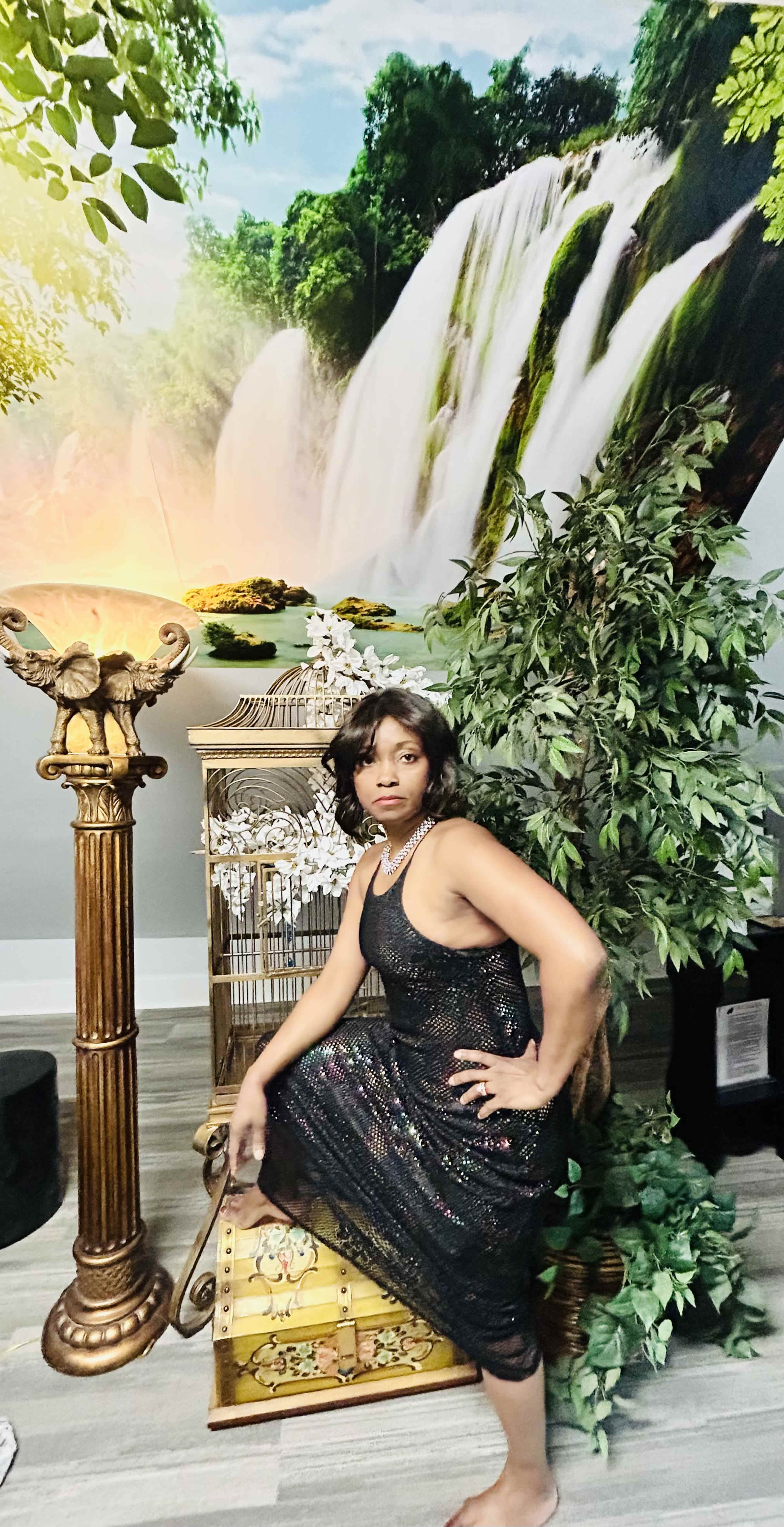 A woman in a sequined dress poses next to a decorative pedestal and lush greenery in front of a scenic mural of a waterfall.