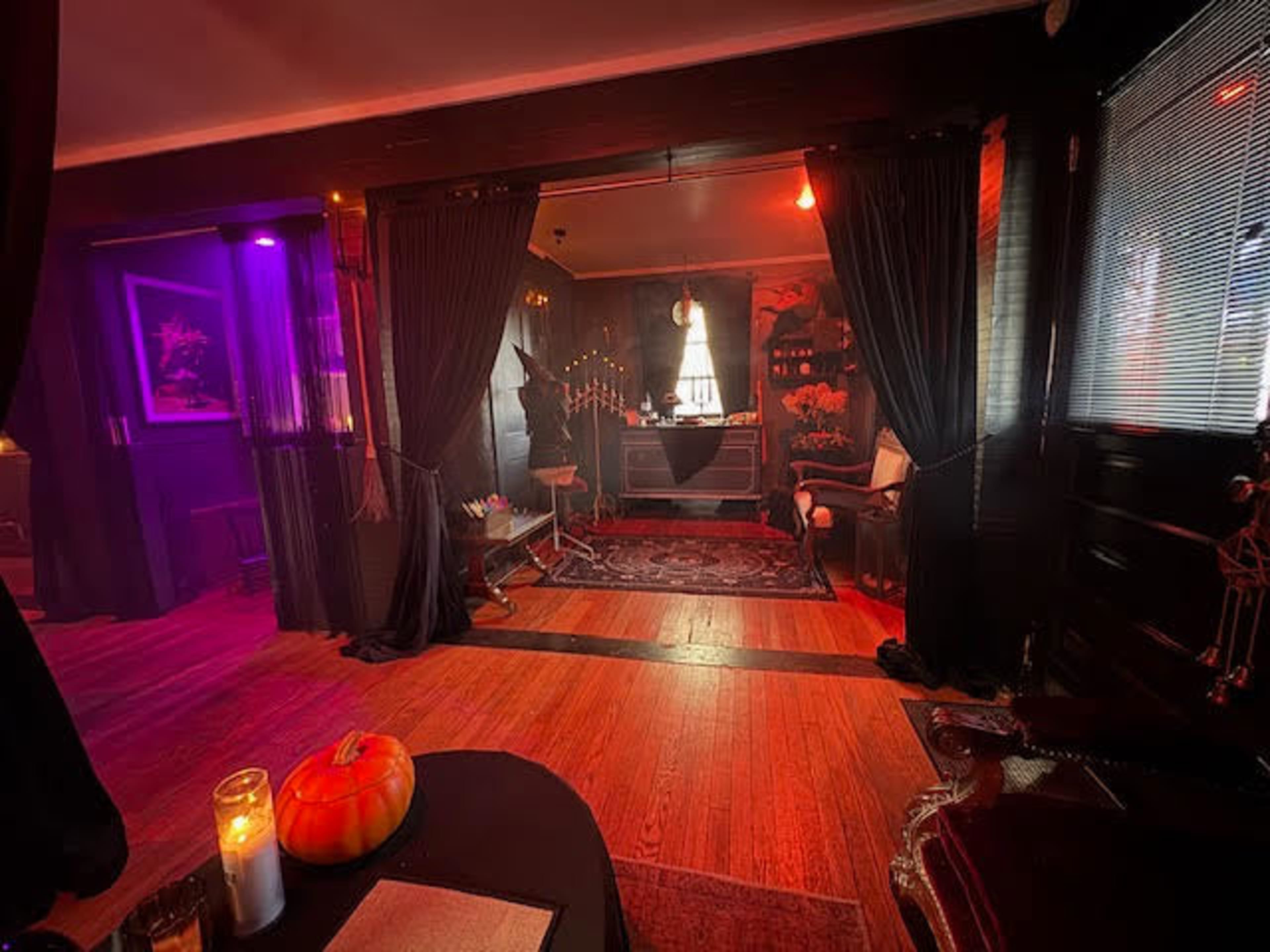 The image shows a dimly lit room decorated for Halloween, featuring a pumpkin, candles, and dark curtains around a central area with furniture and artwork.
