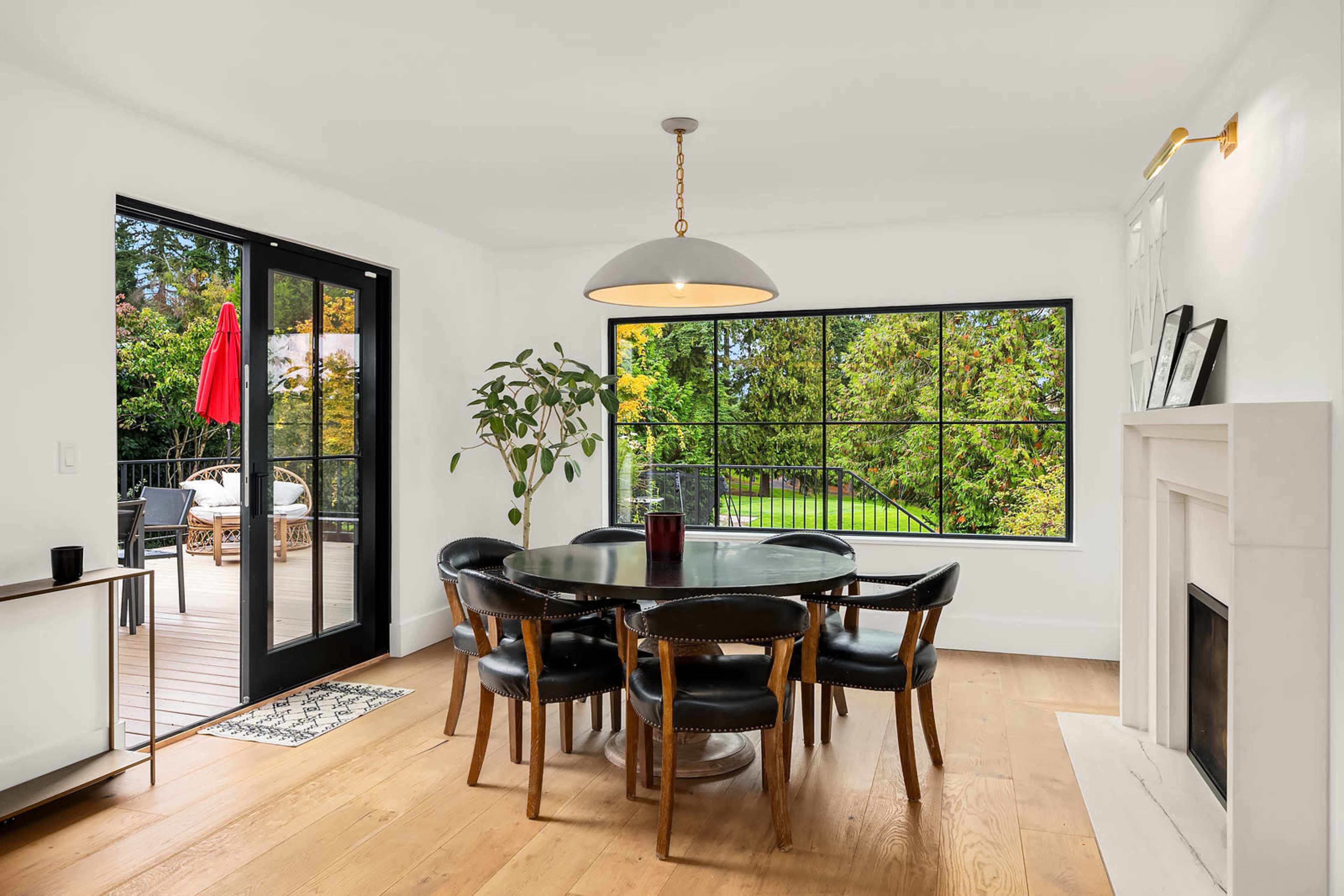 A modern dining area features a round black table surrounded by six chairs, with a view of a deck and garden visible through large windows.