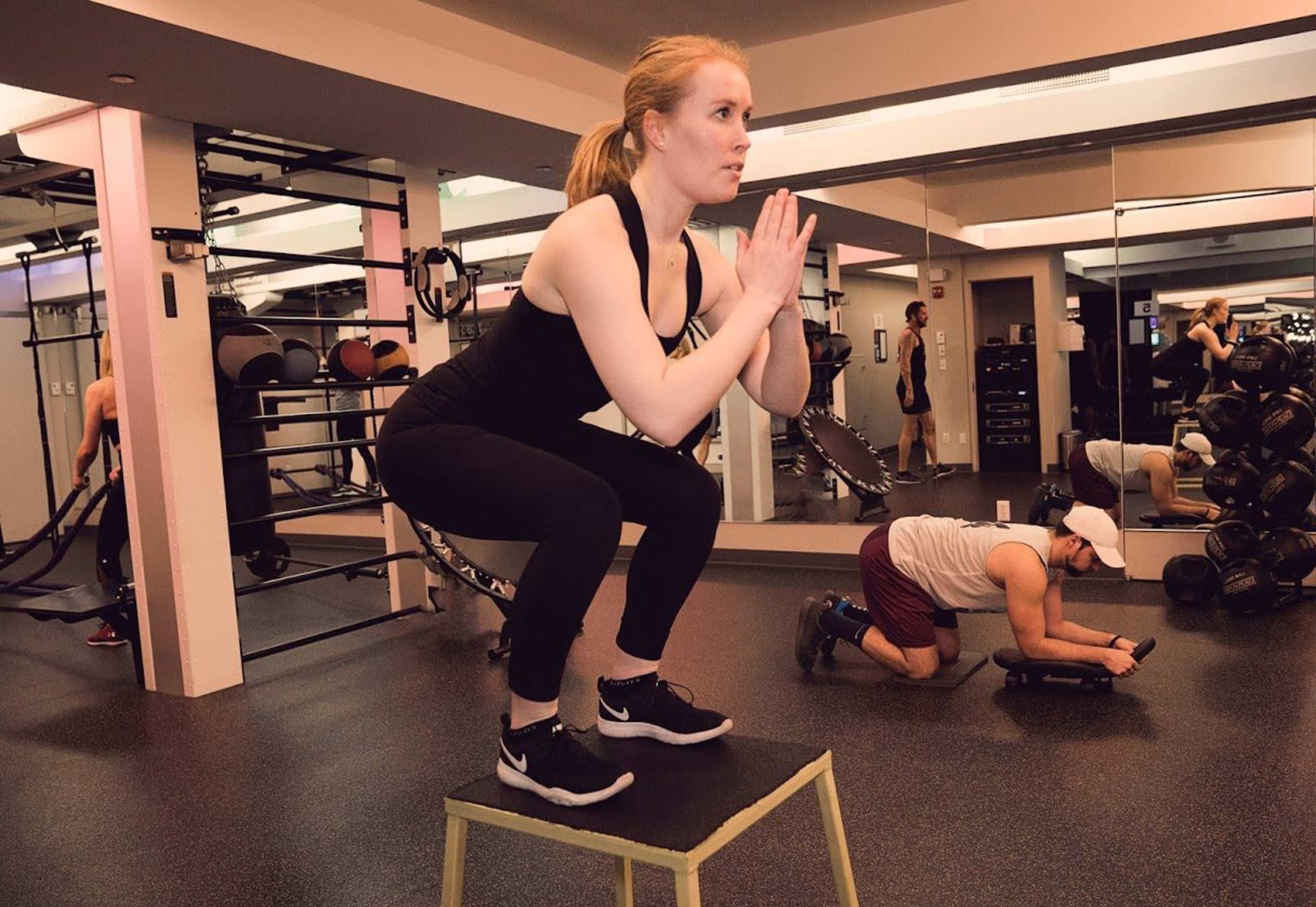A woman performs squats on a box while a man works on the floor with exercise bands in a gym setting.