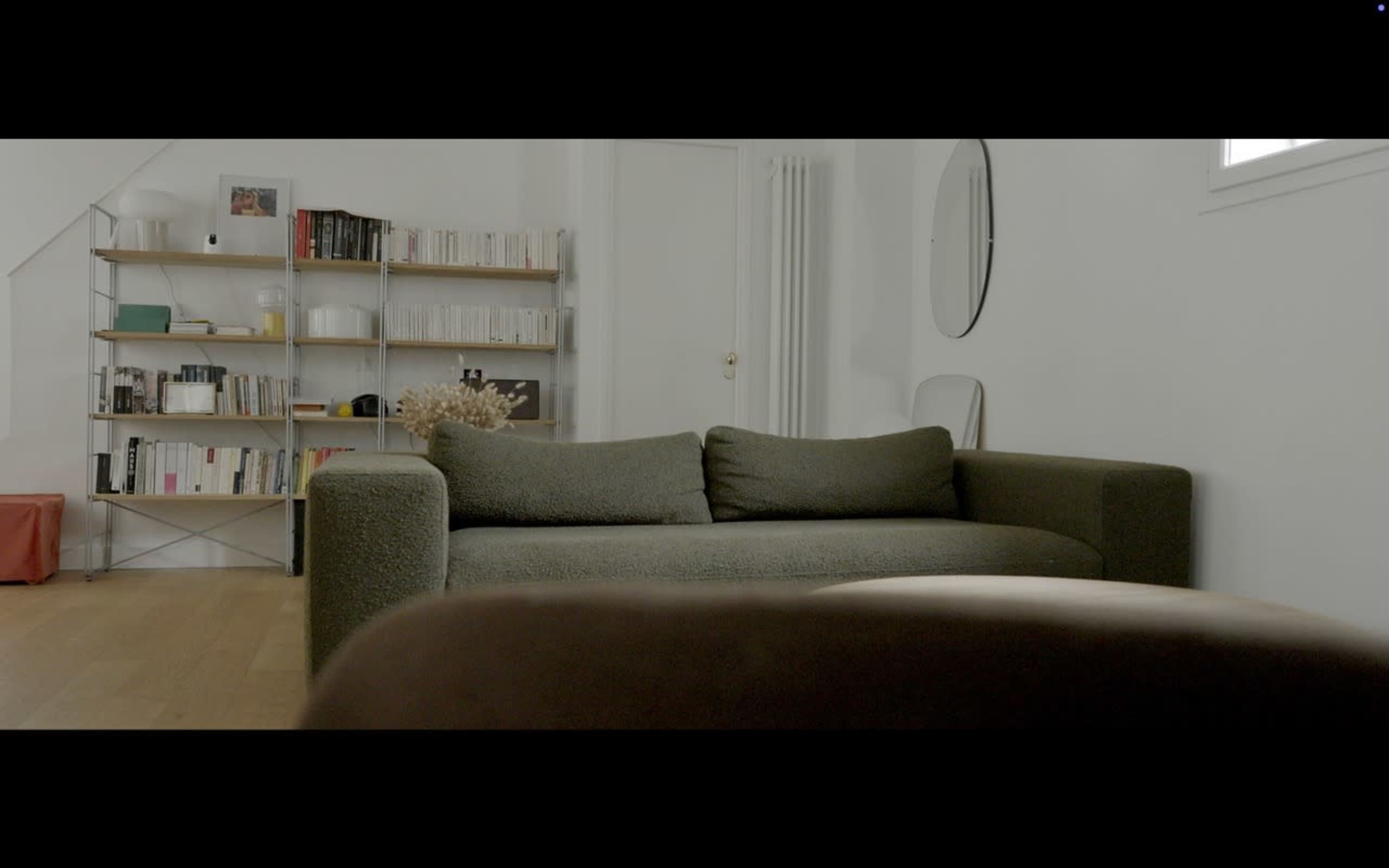 The image shows a modern living room with a green sofa in the foreground, a bookshelf filled with books in the background, and a round mirror hanging on the wall.