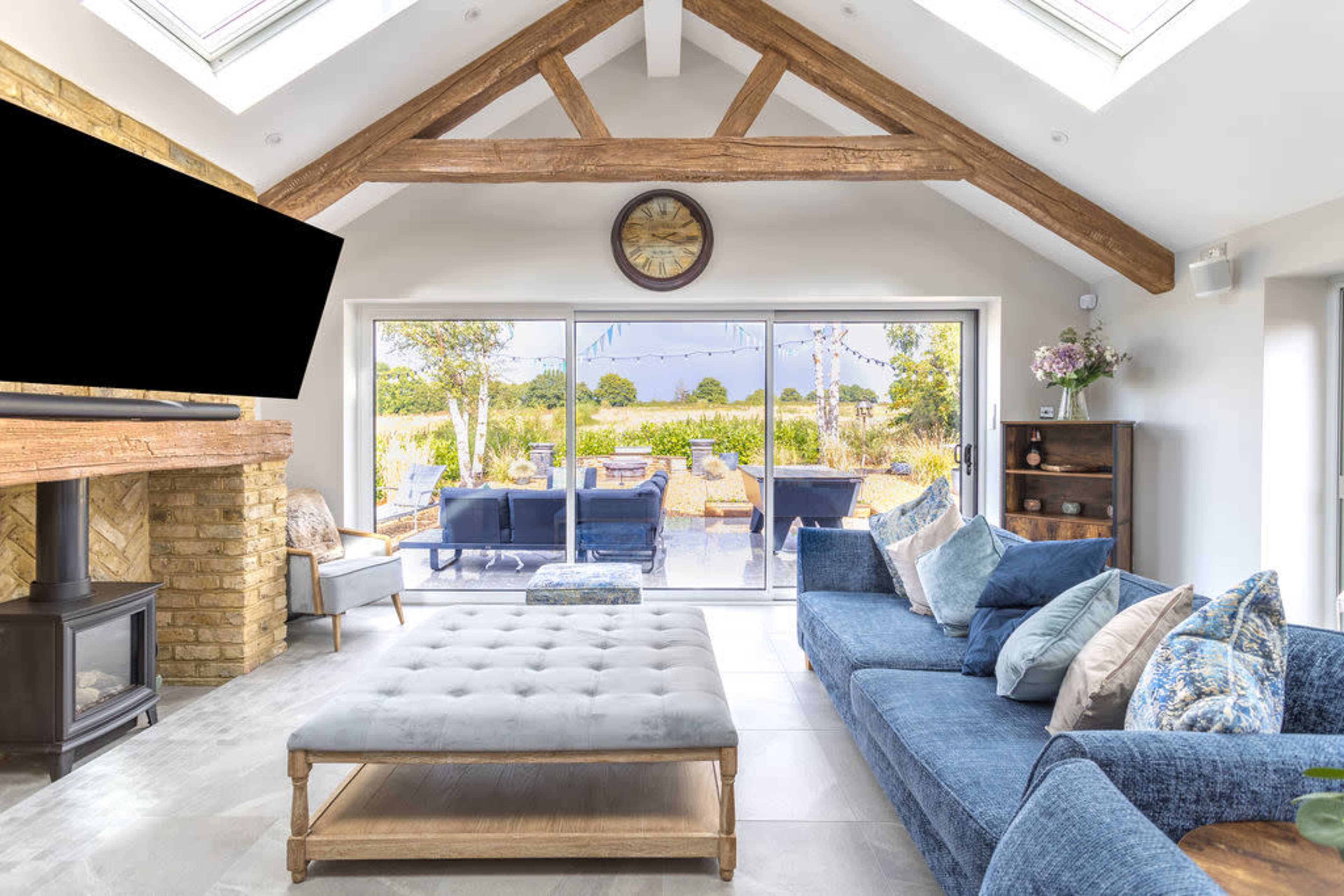 A modern living room features a large sectional sofa, a coffee table, and a flat-screen television, with sliding glass doors opening to a view of an outdoor area with seating and greenery.