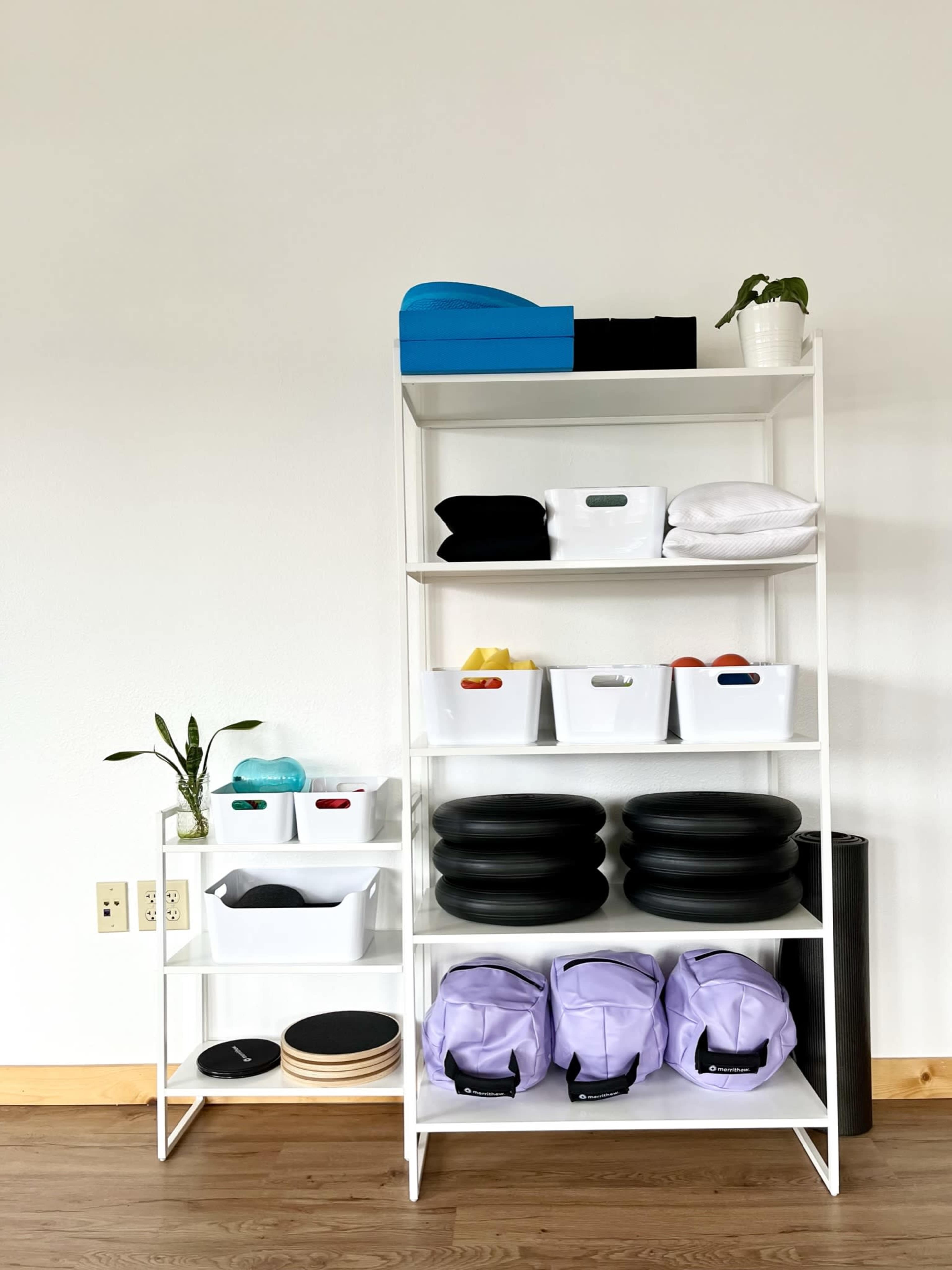 A white shelving unit displays various exercise equipment and storage bins organized neatly against a plain wall.