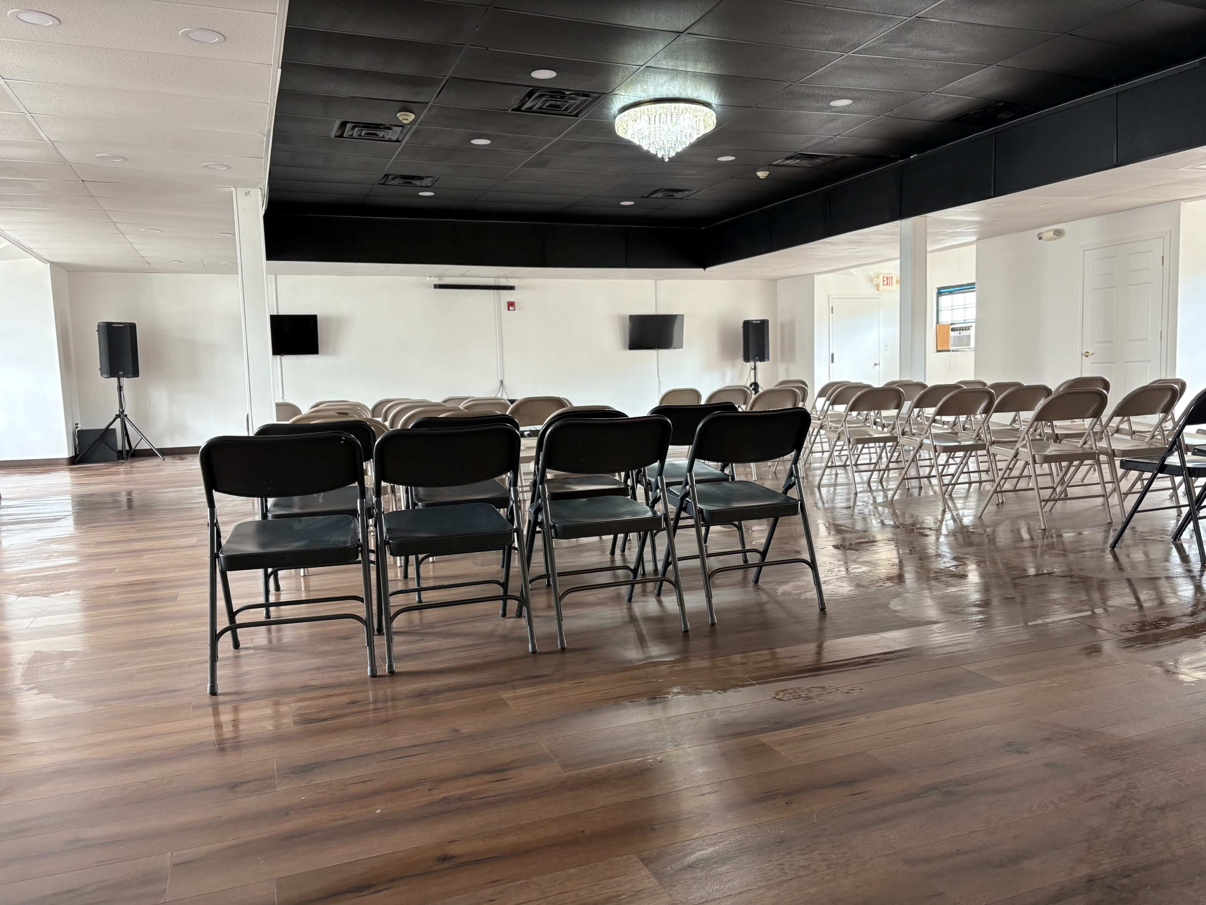 The image shows an empty event space with rows of black chairs arranged on polished wooden flooring, accompanied by two screens on the walls and a chandelier overhead.
