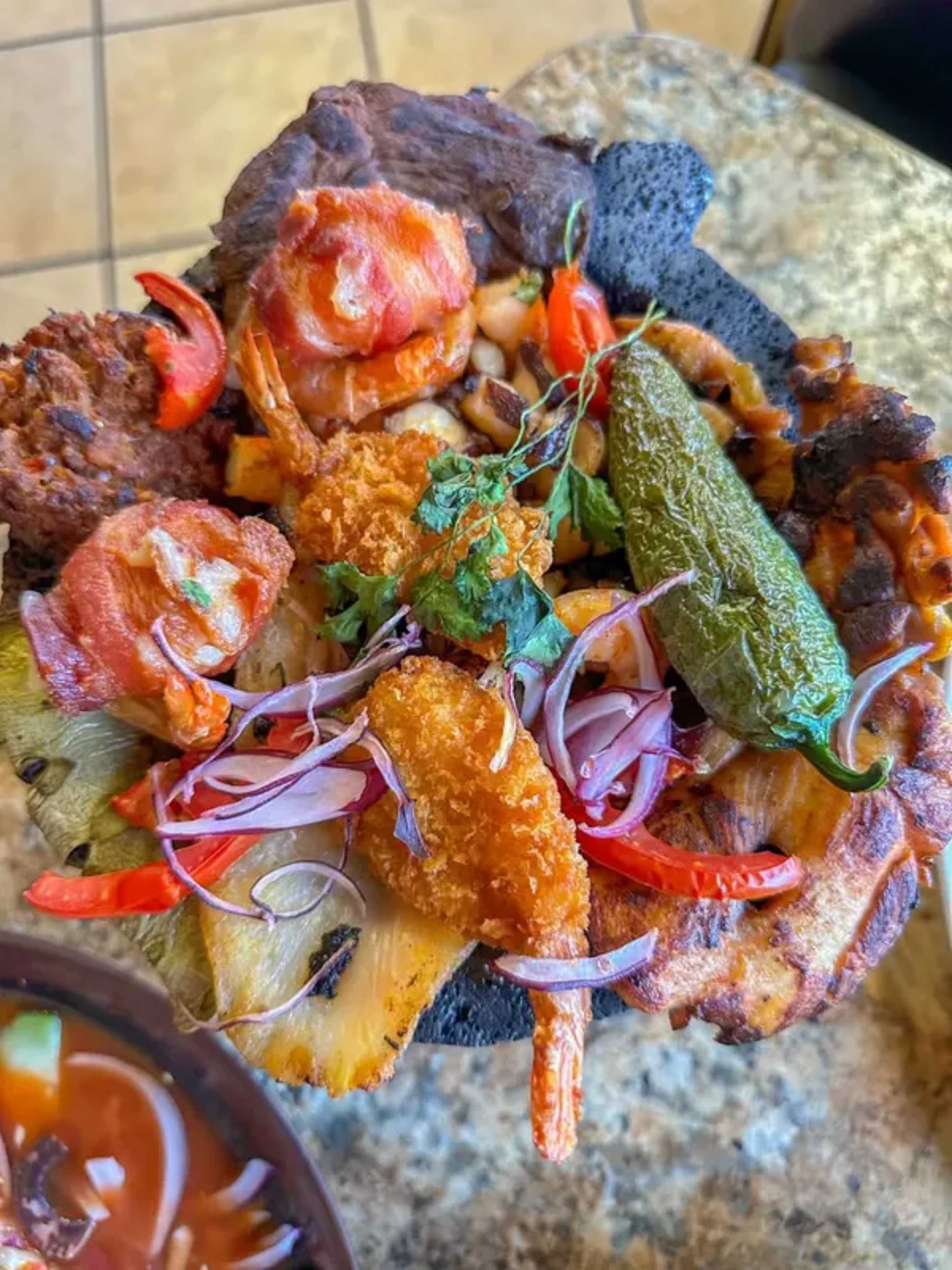 The image shows a colorful platter of grilled meats, shrimp, and vegetables served in a traditional volcanic stone dish, accompanied by a bowl of soup.