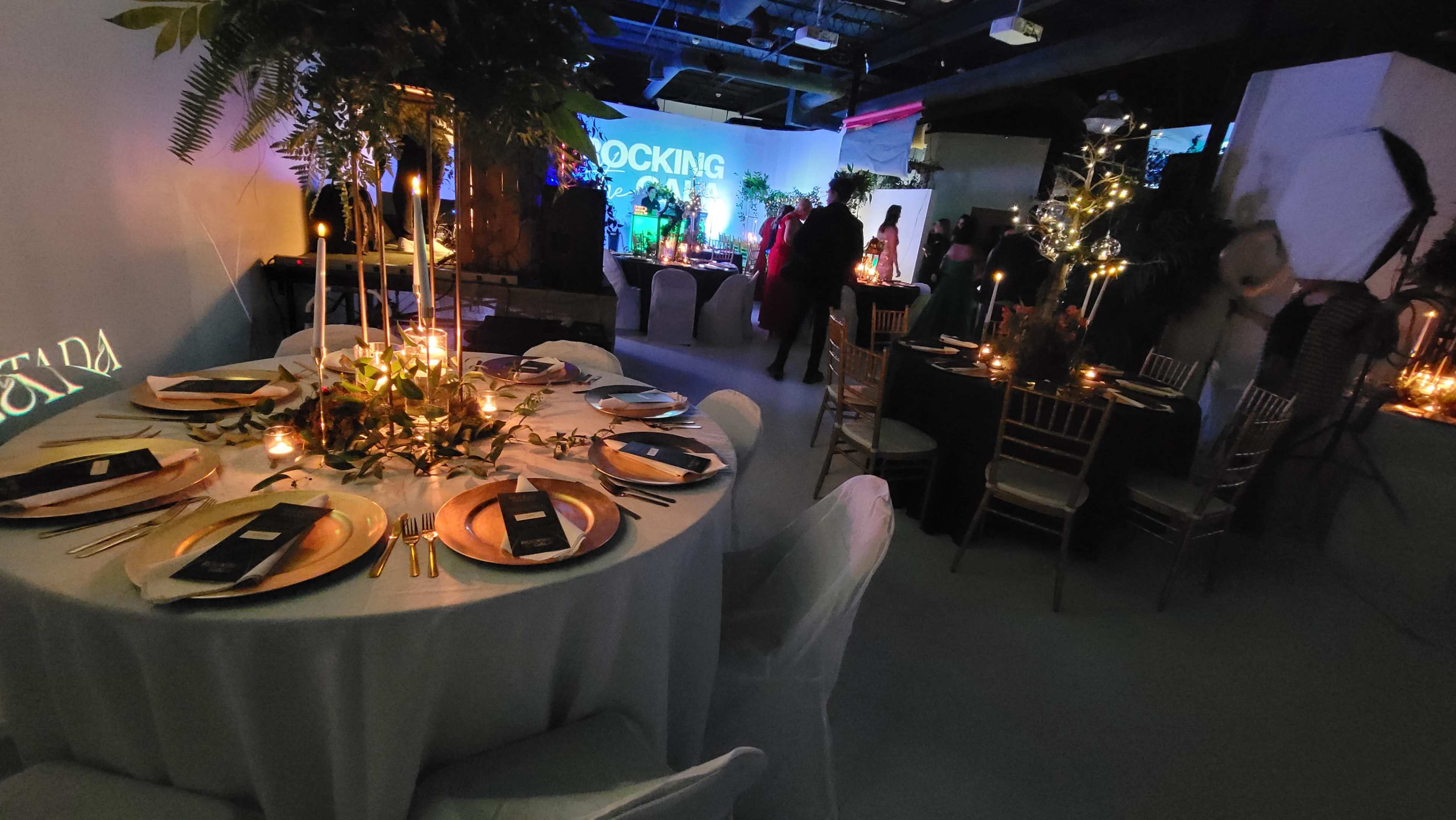 A well-decorated event space features elegantly set tables with candles and greenery, while guests mingle in the background.