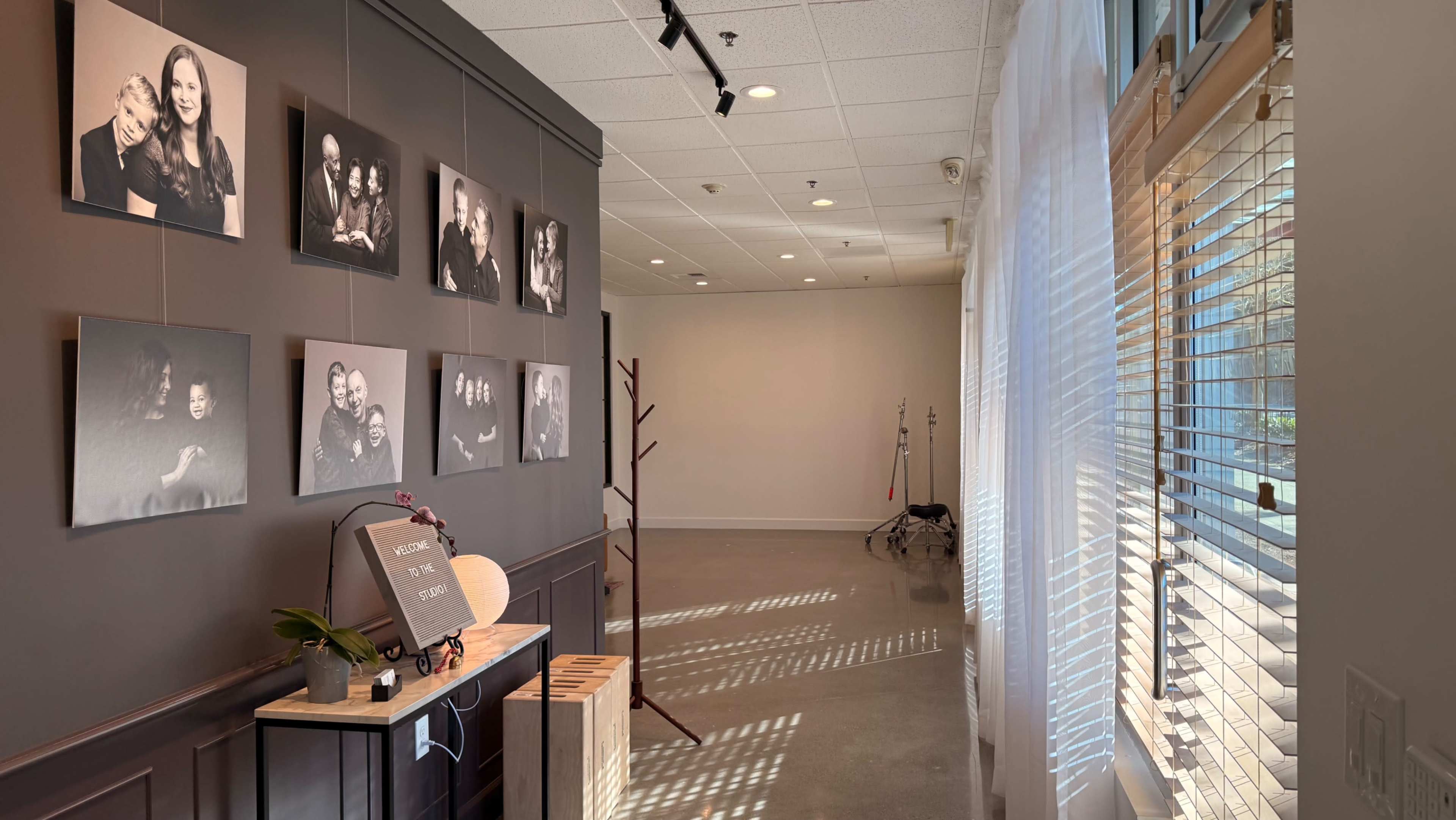 The image shows a well-lit gallery-like space with framed photos on a dark wall and a window on the right casting shadows on the floor.