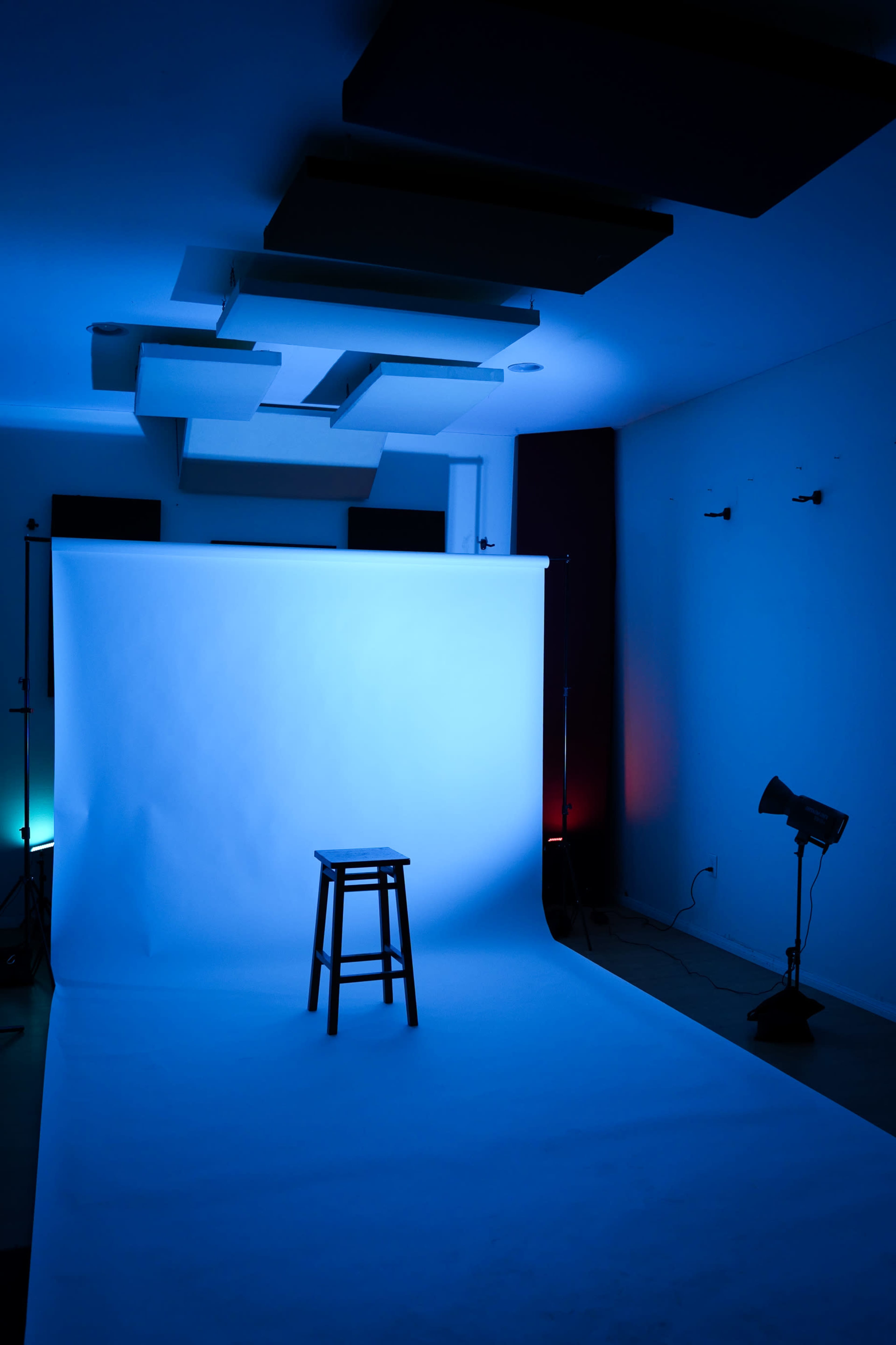 A single wooden stool is positioned in front of a blue backdrop in a studio setting with colored lighting.