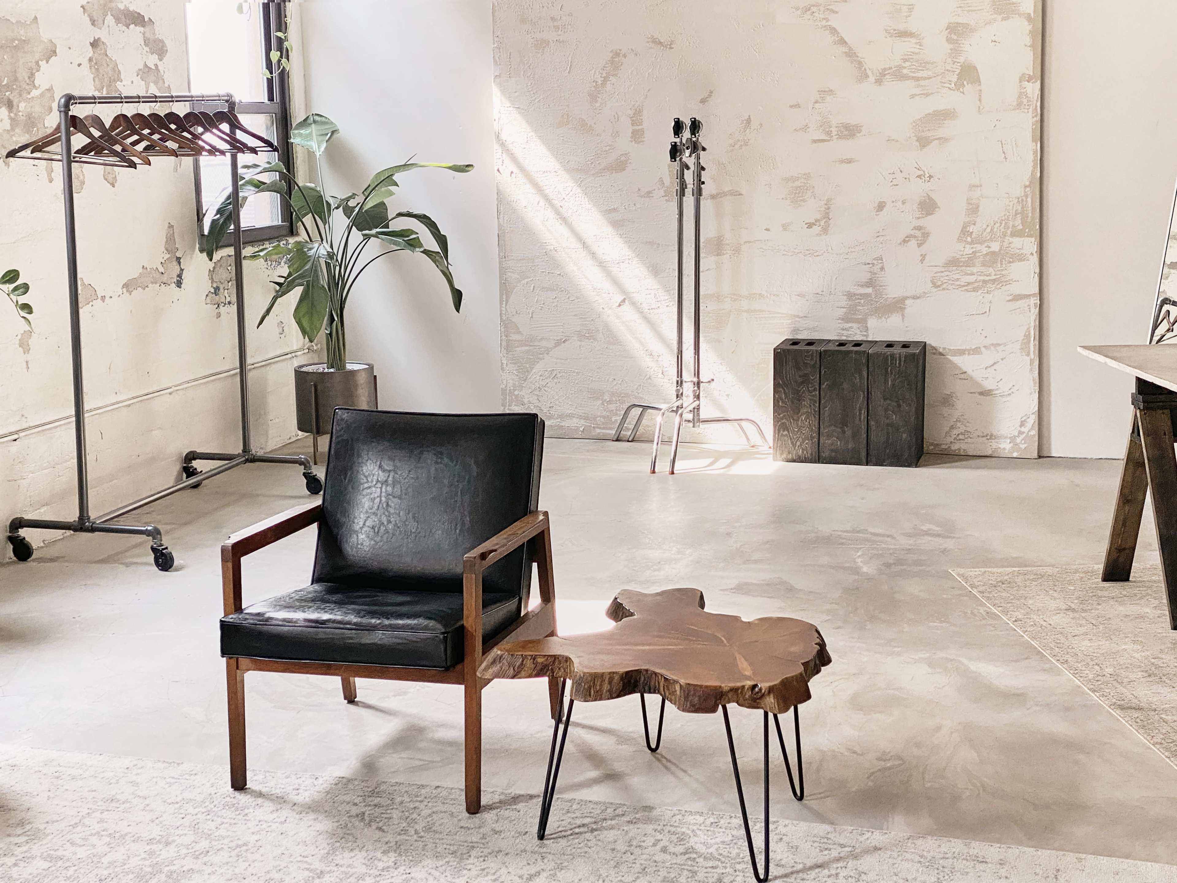 The image shows a minimalist interior space featuring a black chair next to a wooden side table, with clothes racks and a plant in the background.