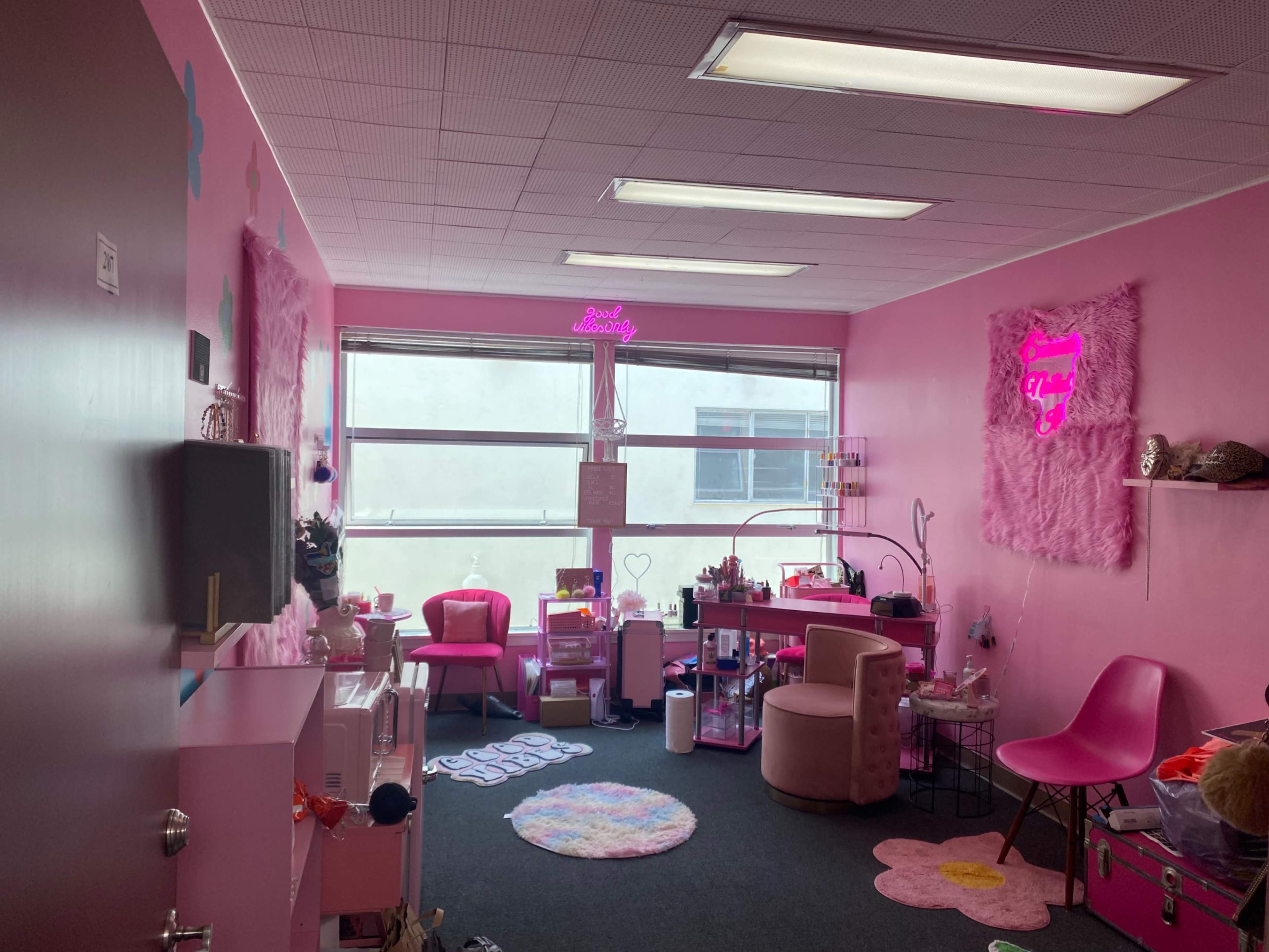 The room is decorated in pink, featuring a seating area with plush chairs, a small table, and a neon sign against the wall, with windows allowing natural light to enter.