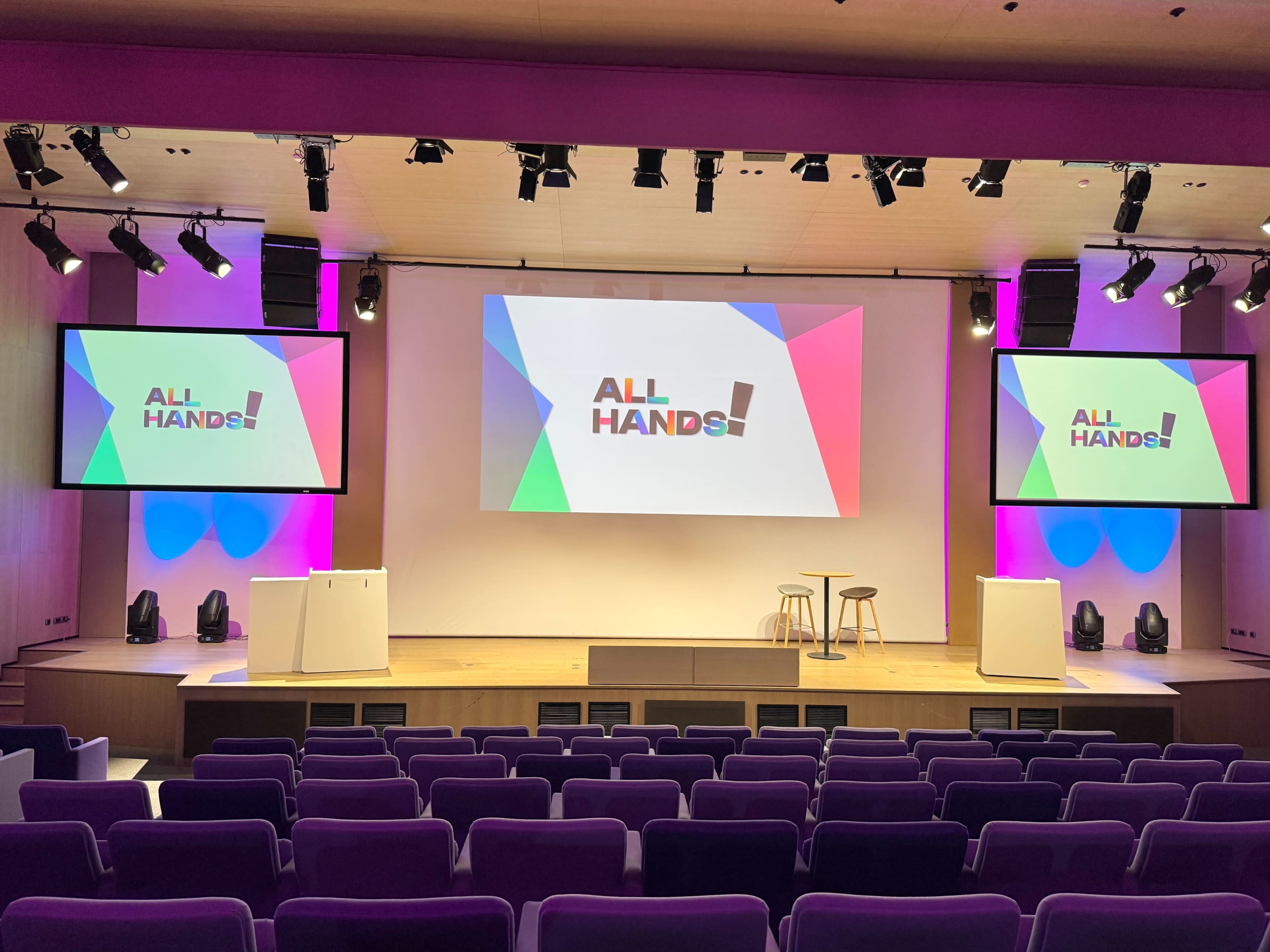 The image shows a stage set up for a presentation, featuring two large screens displaying the text "ALL HANDS!" and a small round table with two stools in front of a seated audience area.