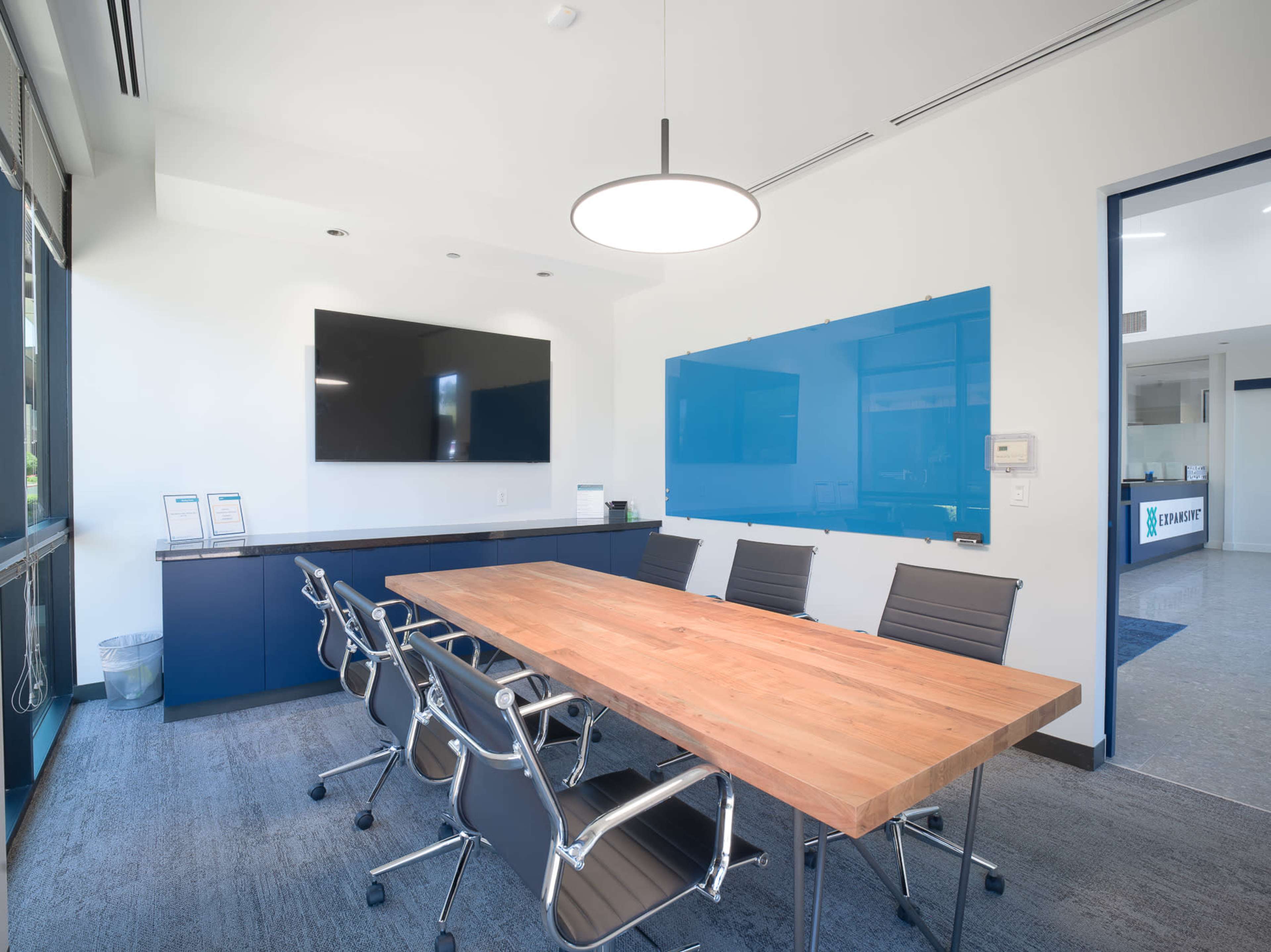 A modern conference room features a large wooden table surrounded by ergonomic chairs, a wall-mounted TV, and a blue glass whiteboard.