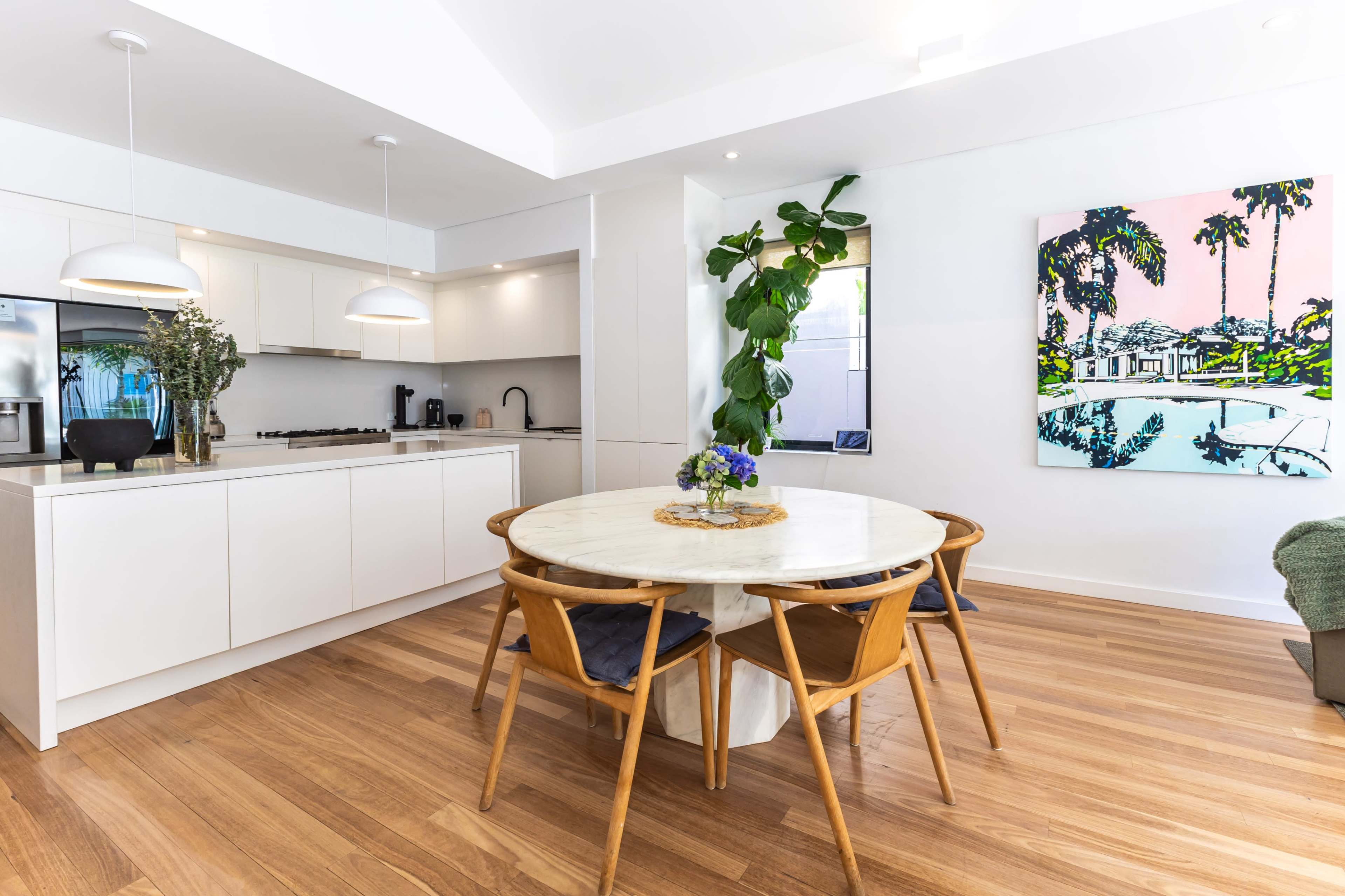 A bright, modern kitchen-dining area features a round marble table surrounded by wooden chairs, with a large window showcasing greenery and a colorful painting on the wall.