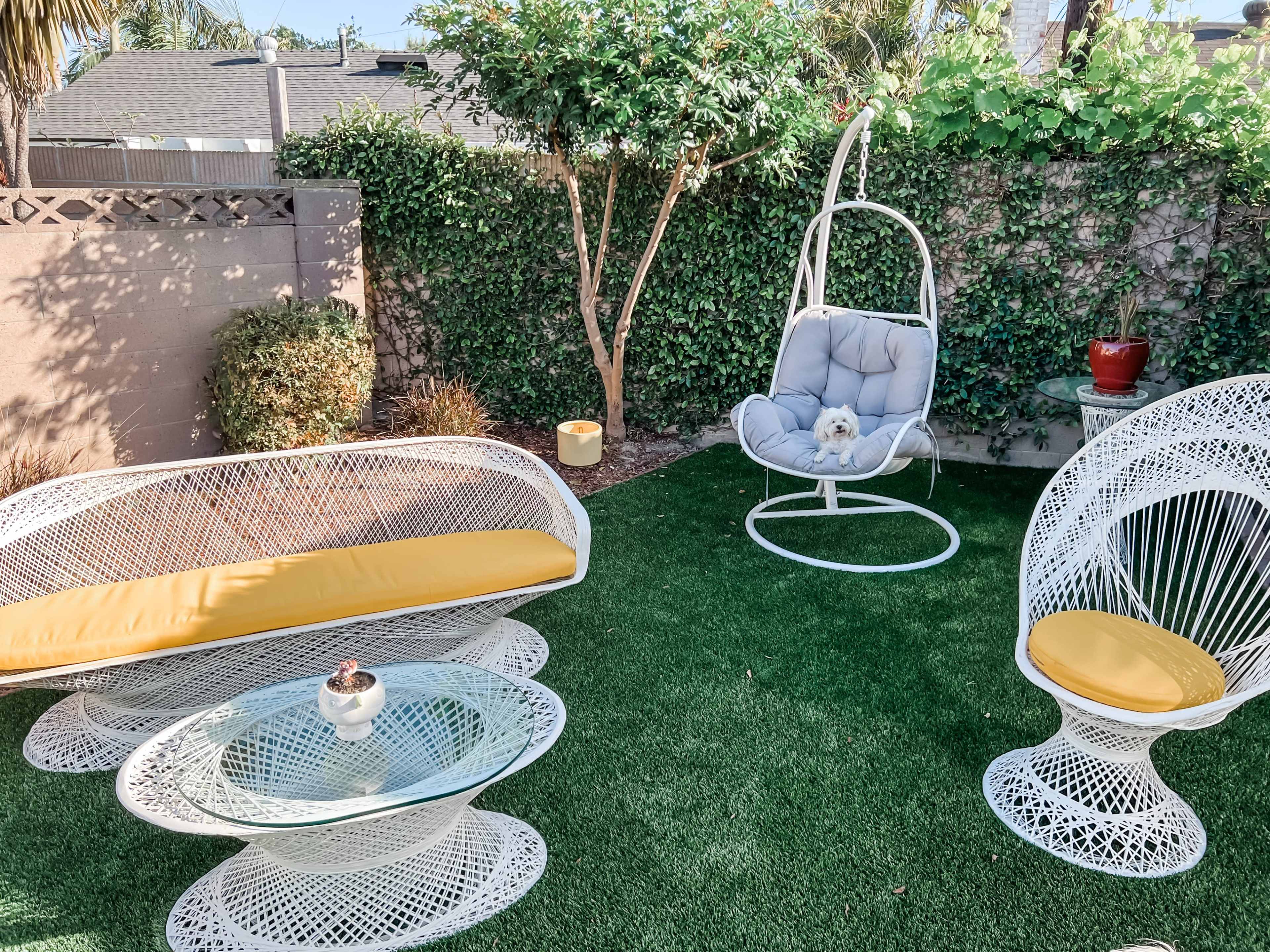 The image shows a small outdoor area with white wicker furniture, including curved chairs and a glass coffee table, surrounded by greenery and a hanging chair with a dog in it.