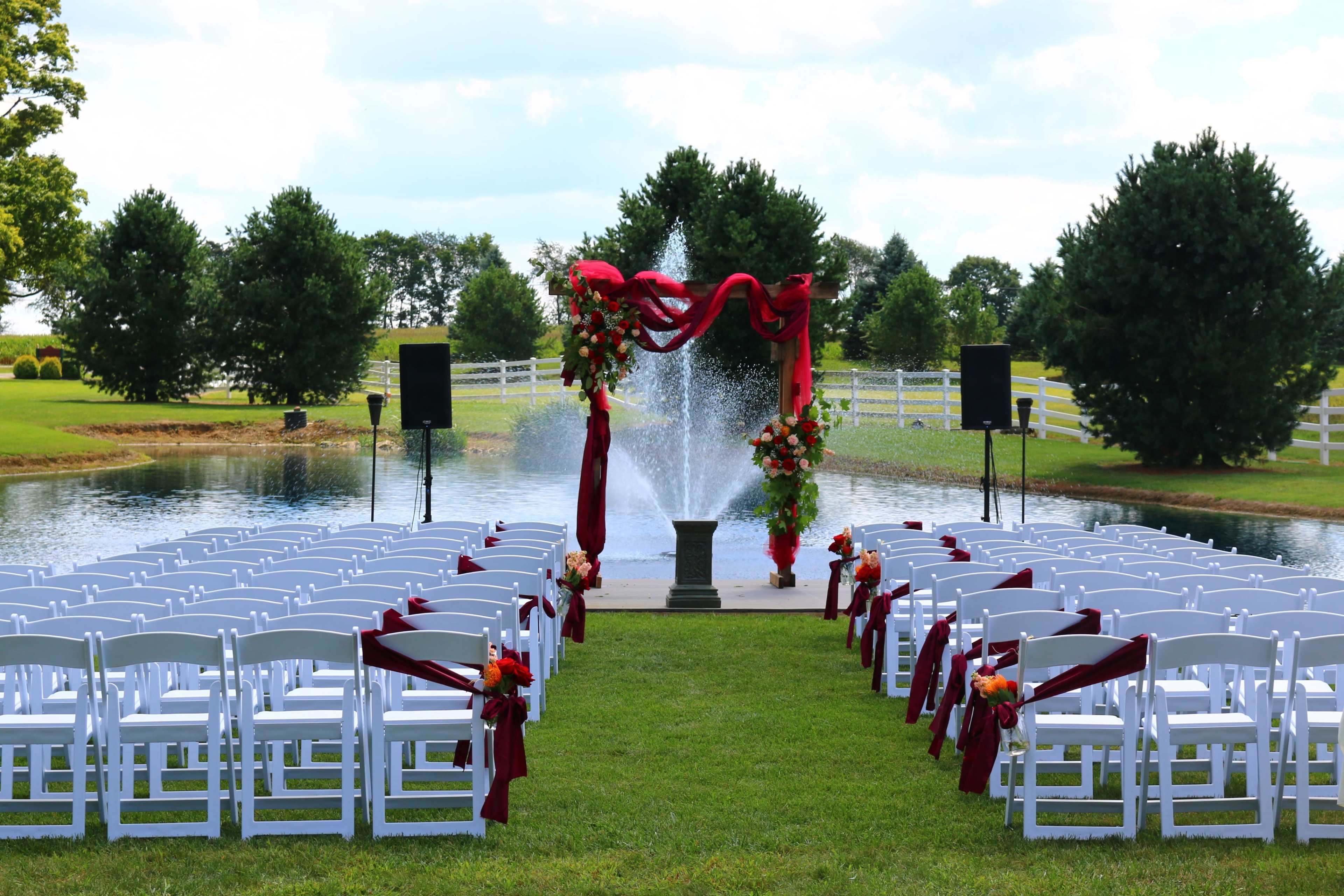 The image shows a wedding ceremony setup with rows of white chairs arranged facing a decorated archway in front of a fountain and a serene pond.