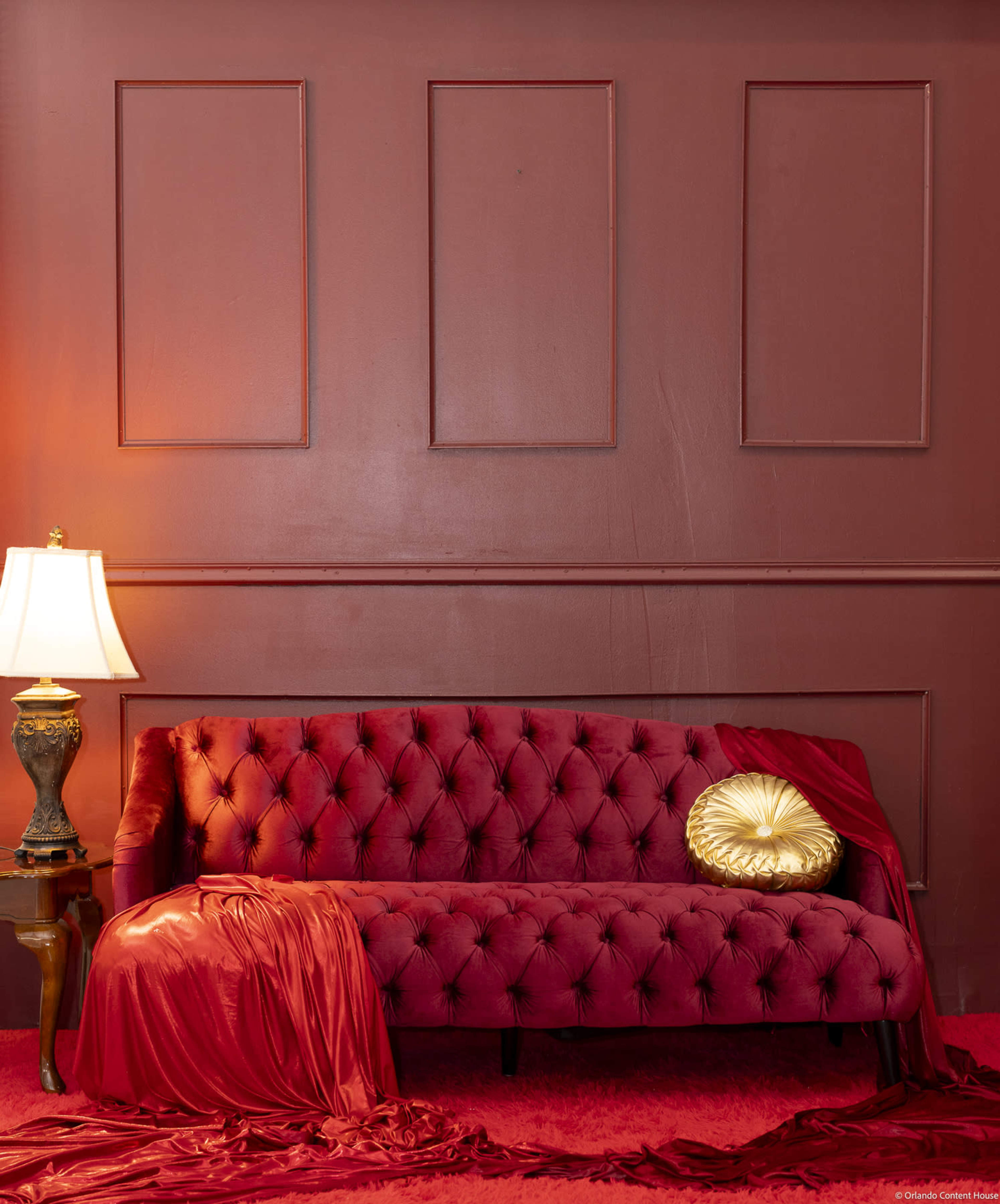 A red velvet sofa is placed against a matching red wall, accompanied by a decorative gold pillow and a lamp on a side table.