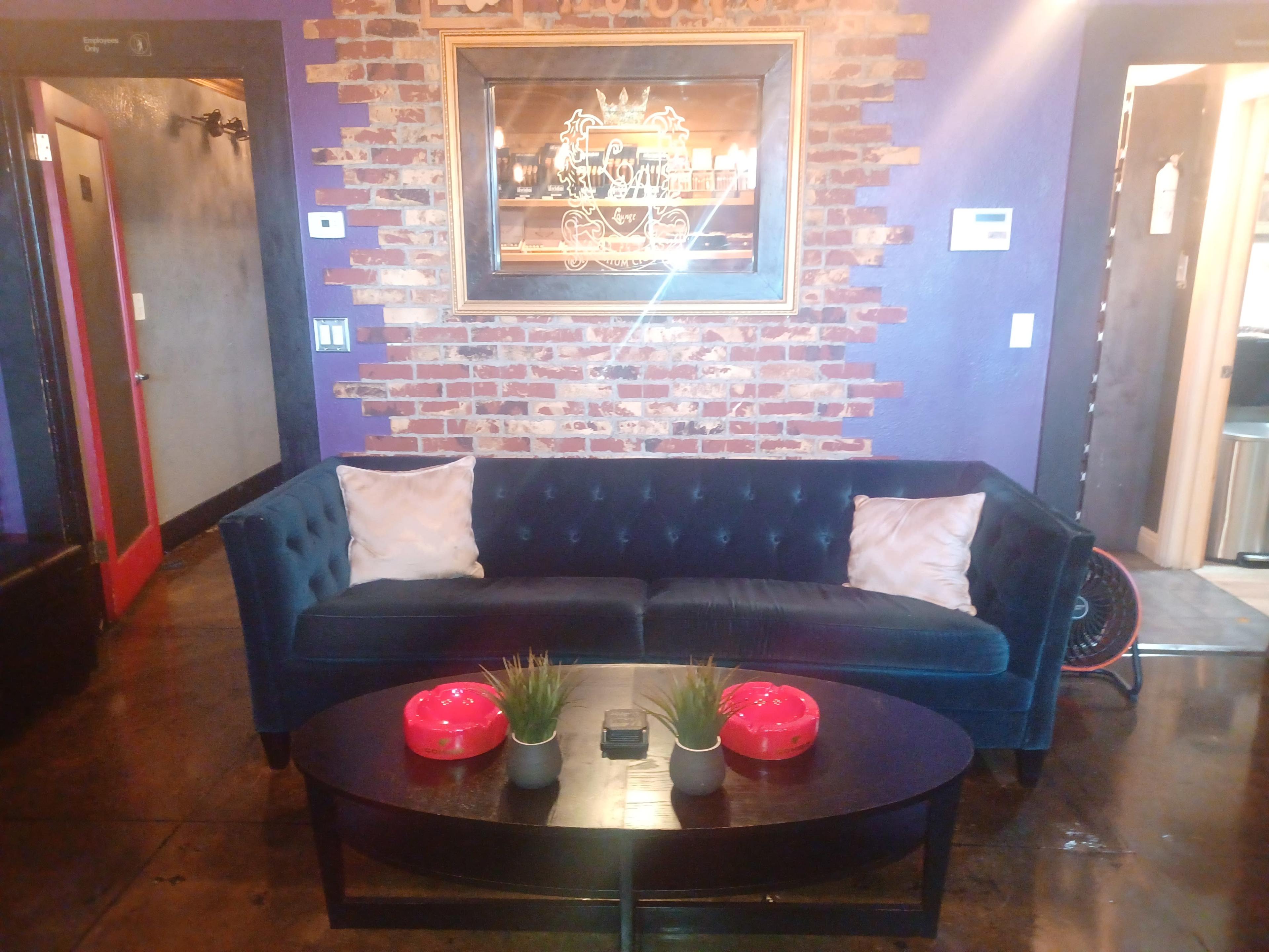 A dark blue velvet sofa with two decorative pillows facing a round black coffee table, set against a brick wall with framed artwork above.