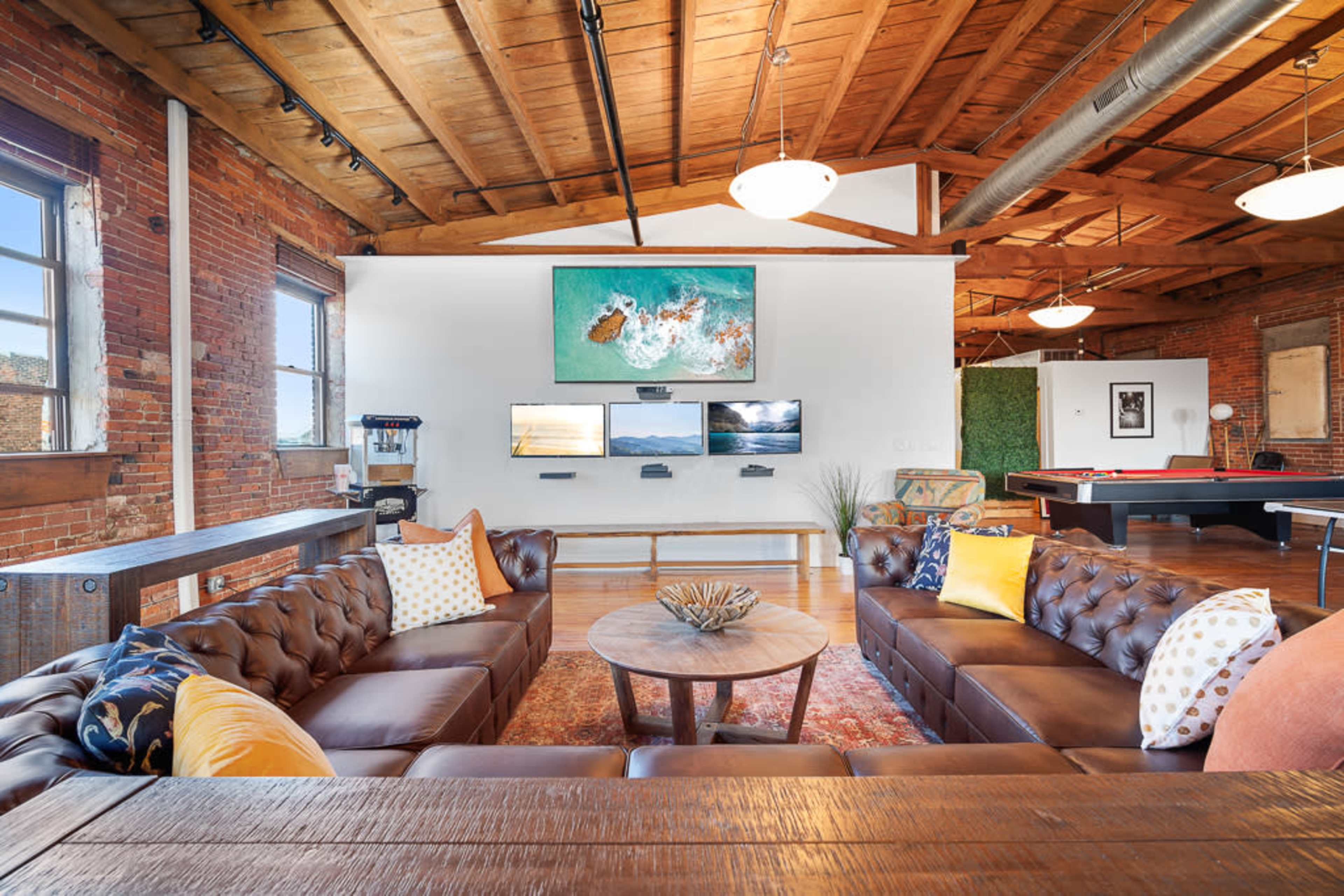A spacious industrial-style living area with exposed brick walls, wooden beams, and two brown leather sofas arranged around a round coffee table, with multiple screens mounted on the wall and a pool table in the background.