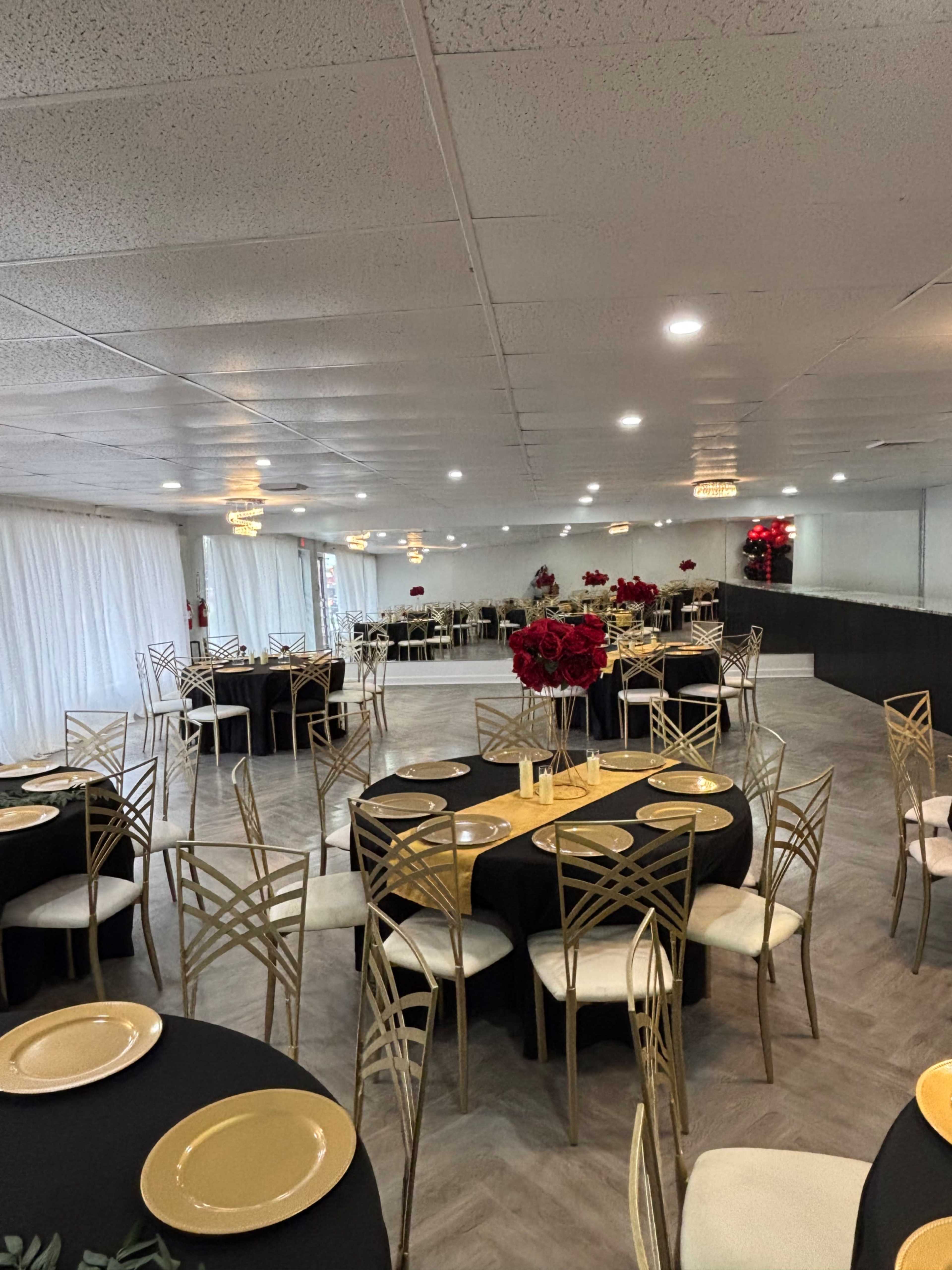The image shows a banquet hall set up with round tables covered in black tablecloths, gold plates, and decorated with red floral arrangements.