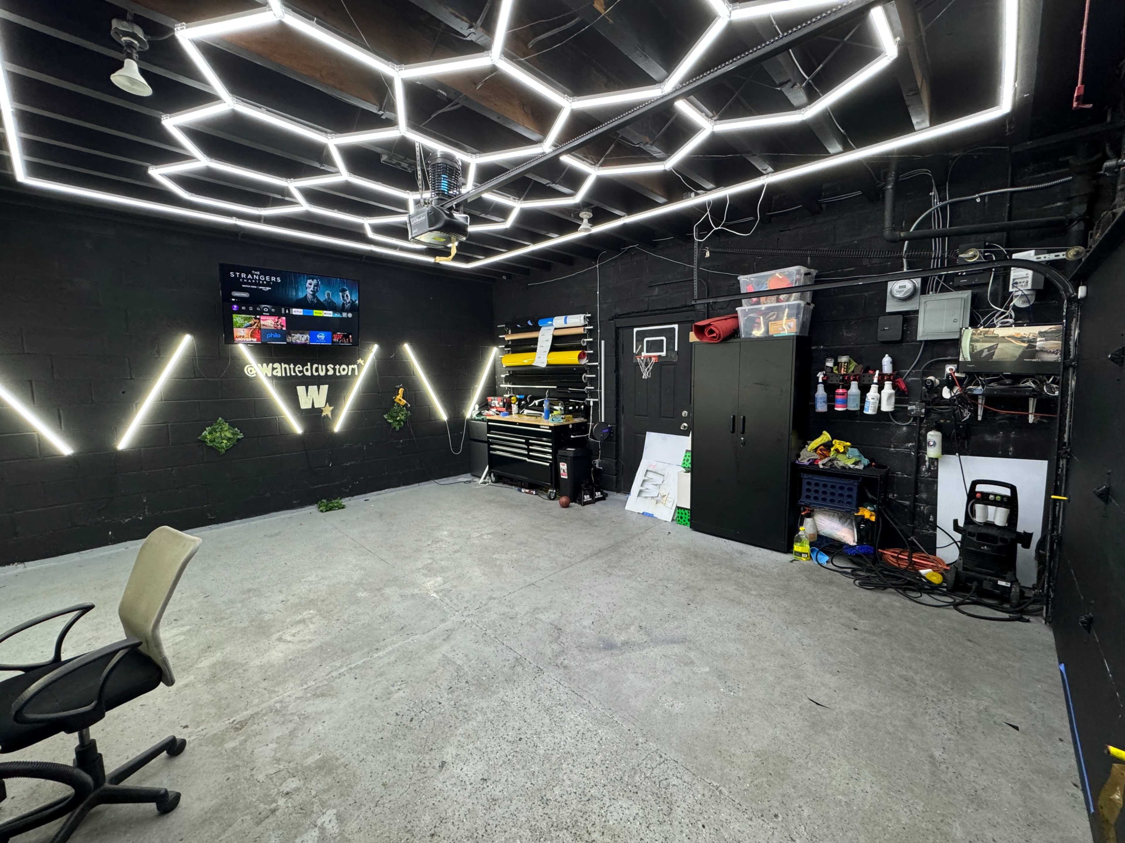 A garage workspace equipped with modern lighting, a wall-mounted TV, and organized tools and supplies on shelves.