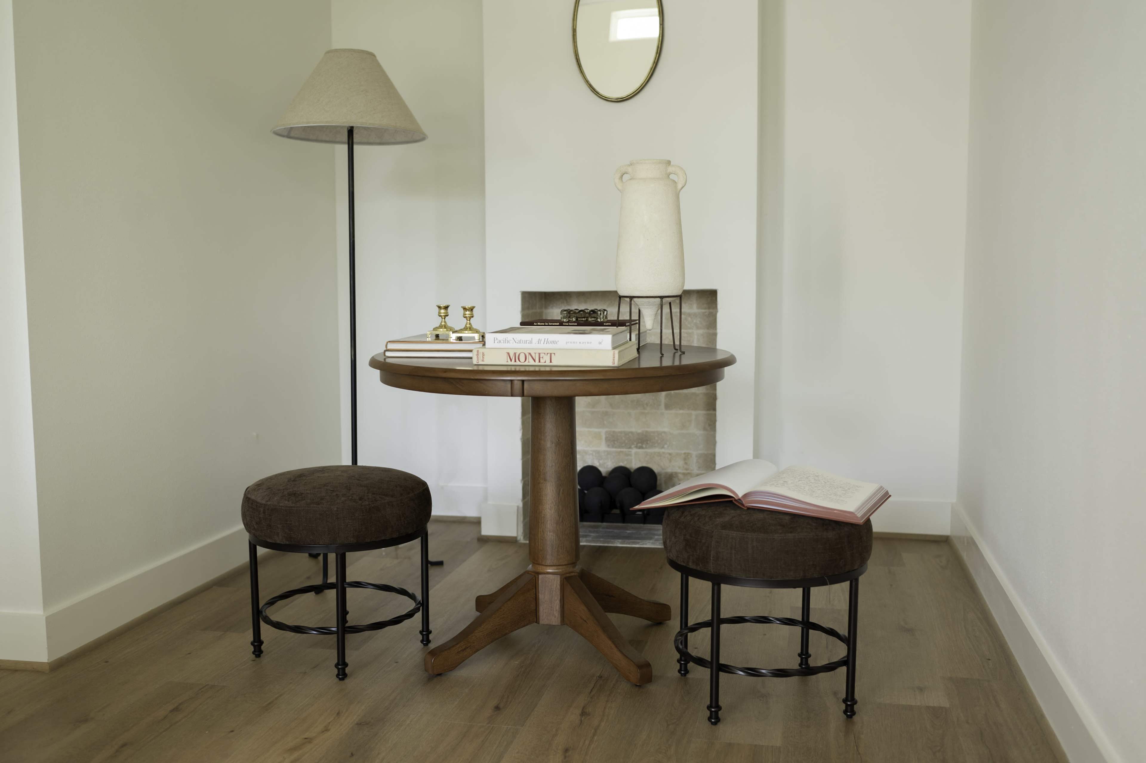 A round wooden table with two upholstered stools is placed beside a floor lamp and a decorative vase, near a minimalist fireplace.