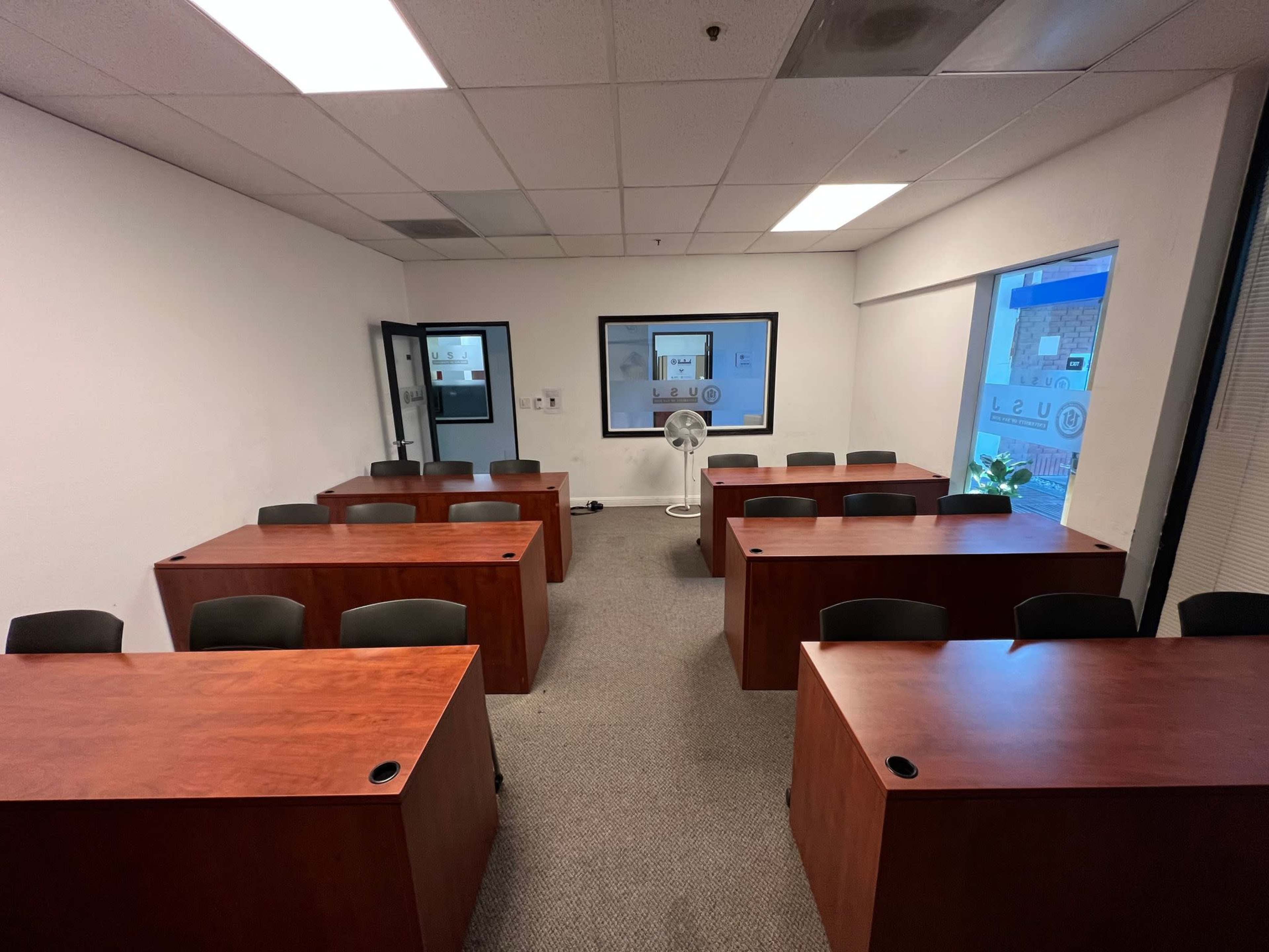 A small classroom with several wooden desks arranged in two rows facing a whiteboard, with a fan and a plant near the window.