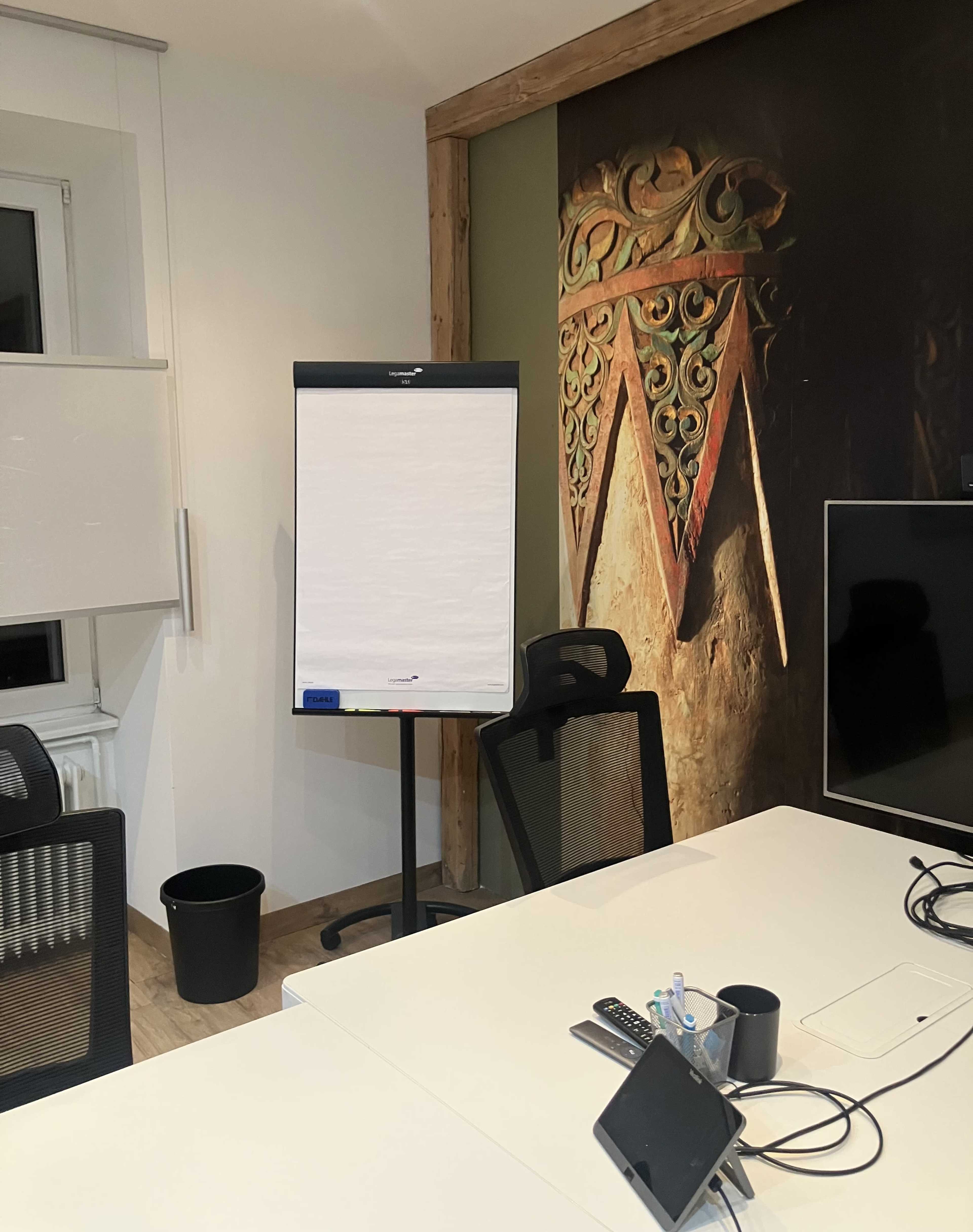 The image shows a conference room with a flip chart on a stand, a black trash bin, and a computer monitor on the table.