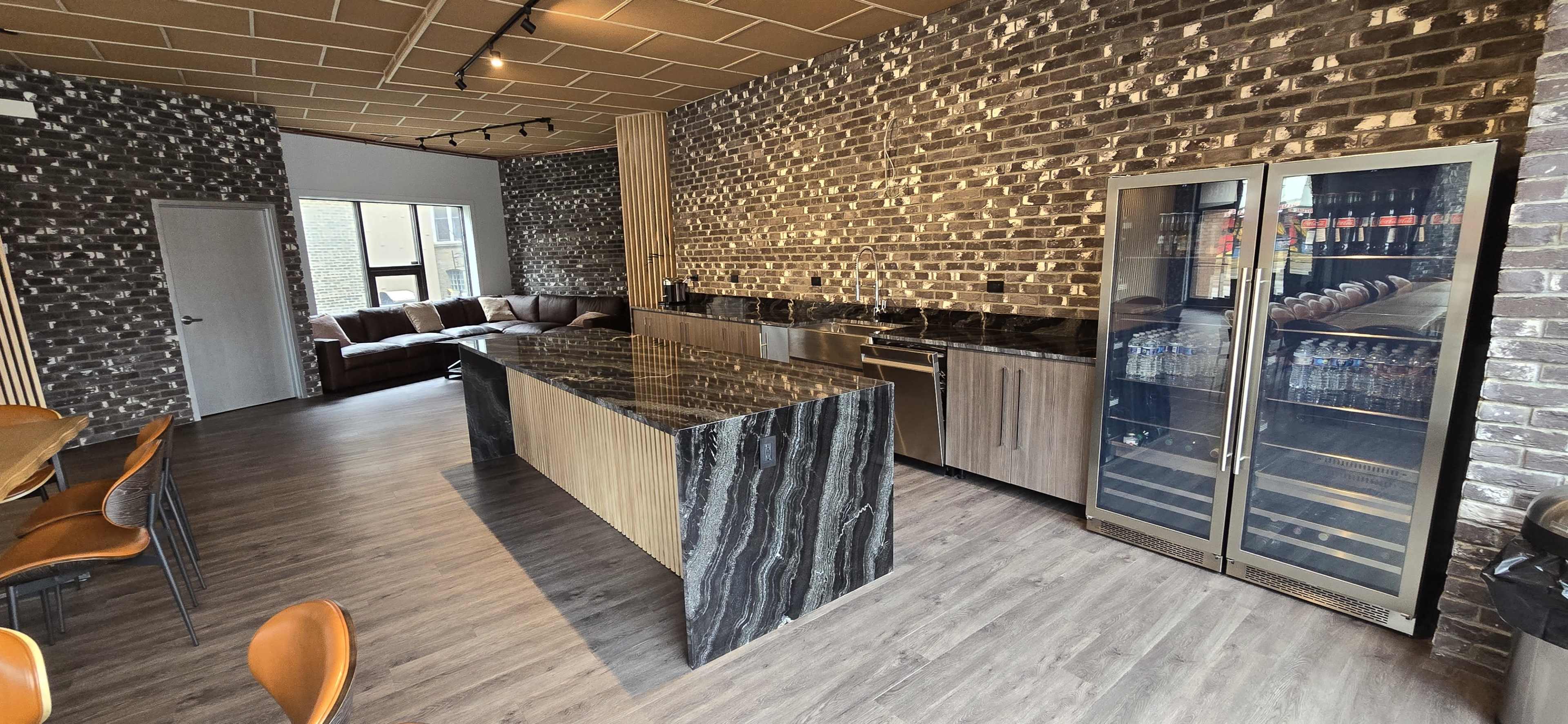 A modern kitchen with a black granite island, stainless steel appliances, and a cozy seating area featuring a brown couch against a brick wall backdrop.