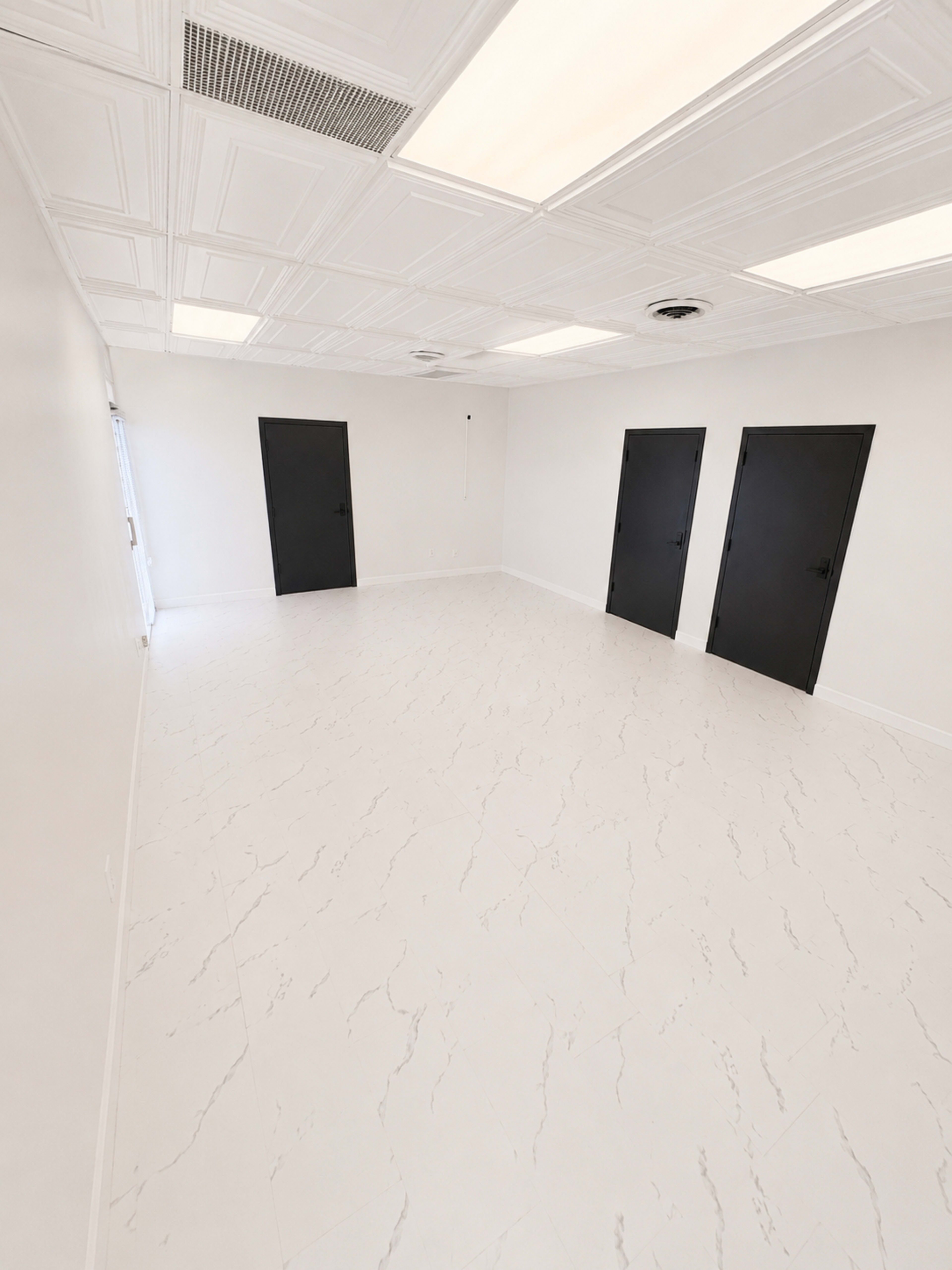 The image shows a spacious, empty room with white walls, a light-colored floor, and three black doors on one side.