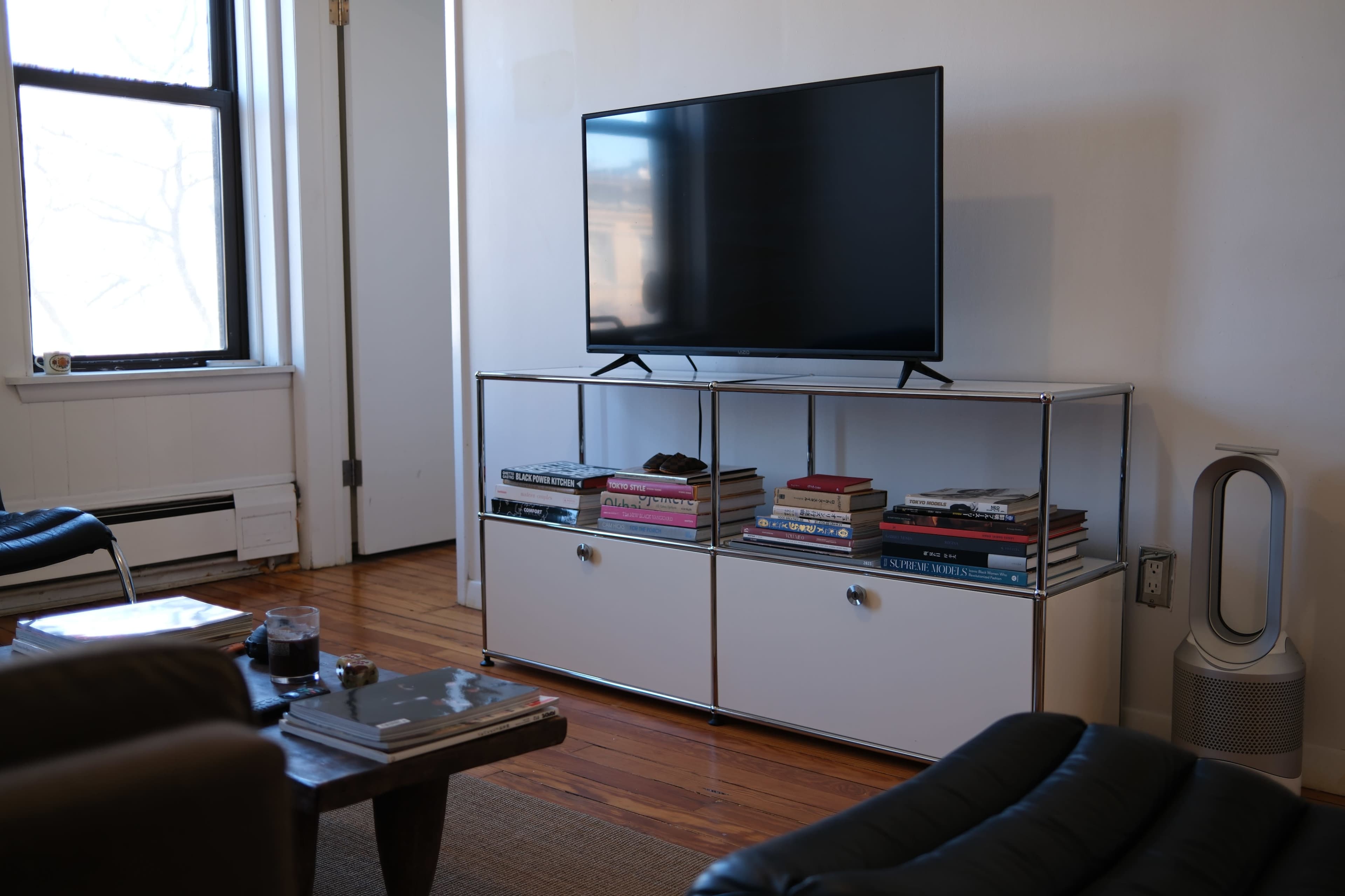 A television sits on a modern stand with open shelves filled with books, next to a black chair and a white wall.