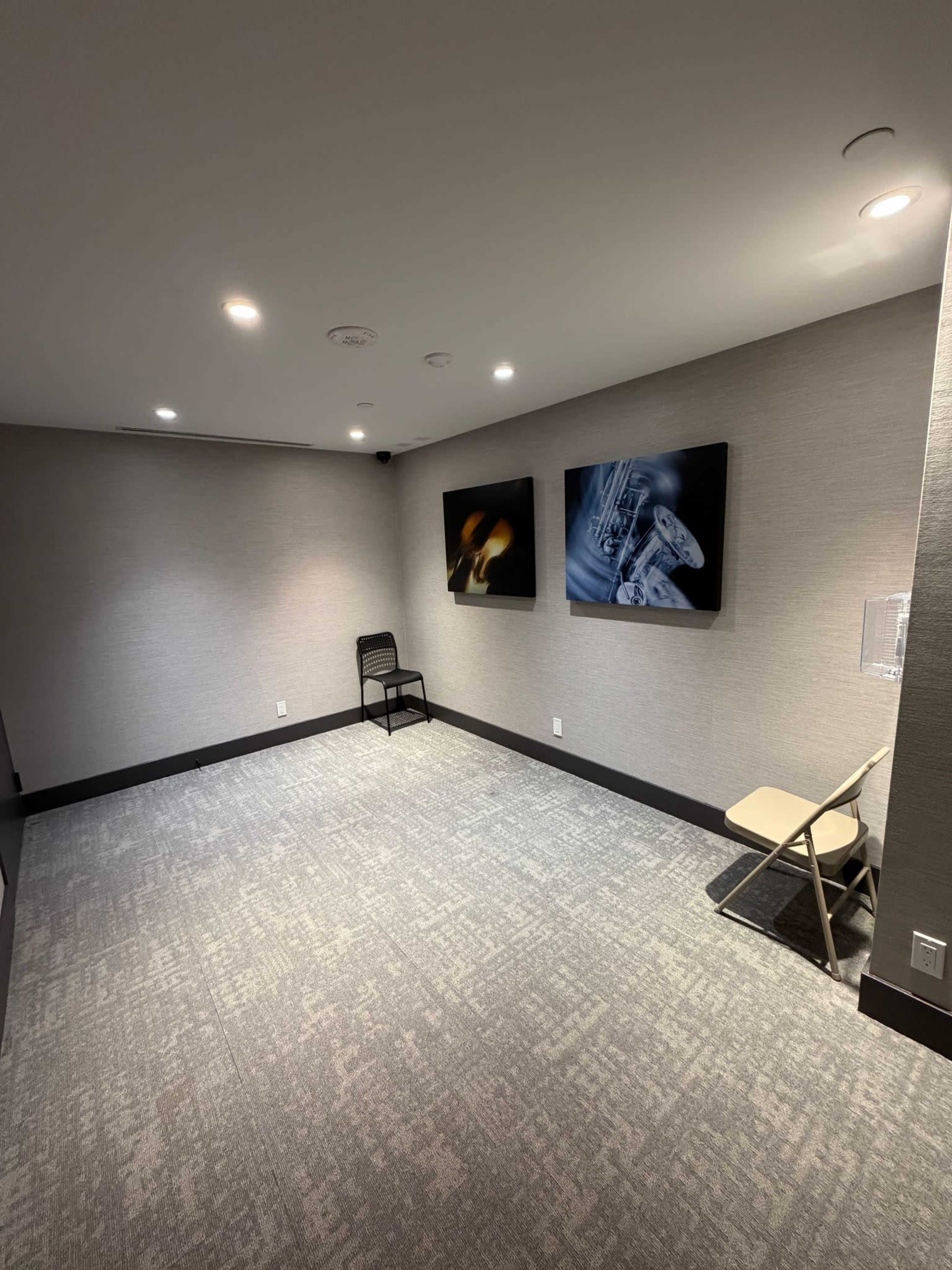 The image shows a sparsely furnished room with two framed artworks on the wall, a black chair, and a folding chair on a carpeted floor.