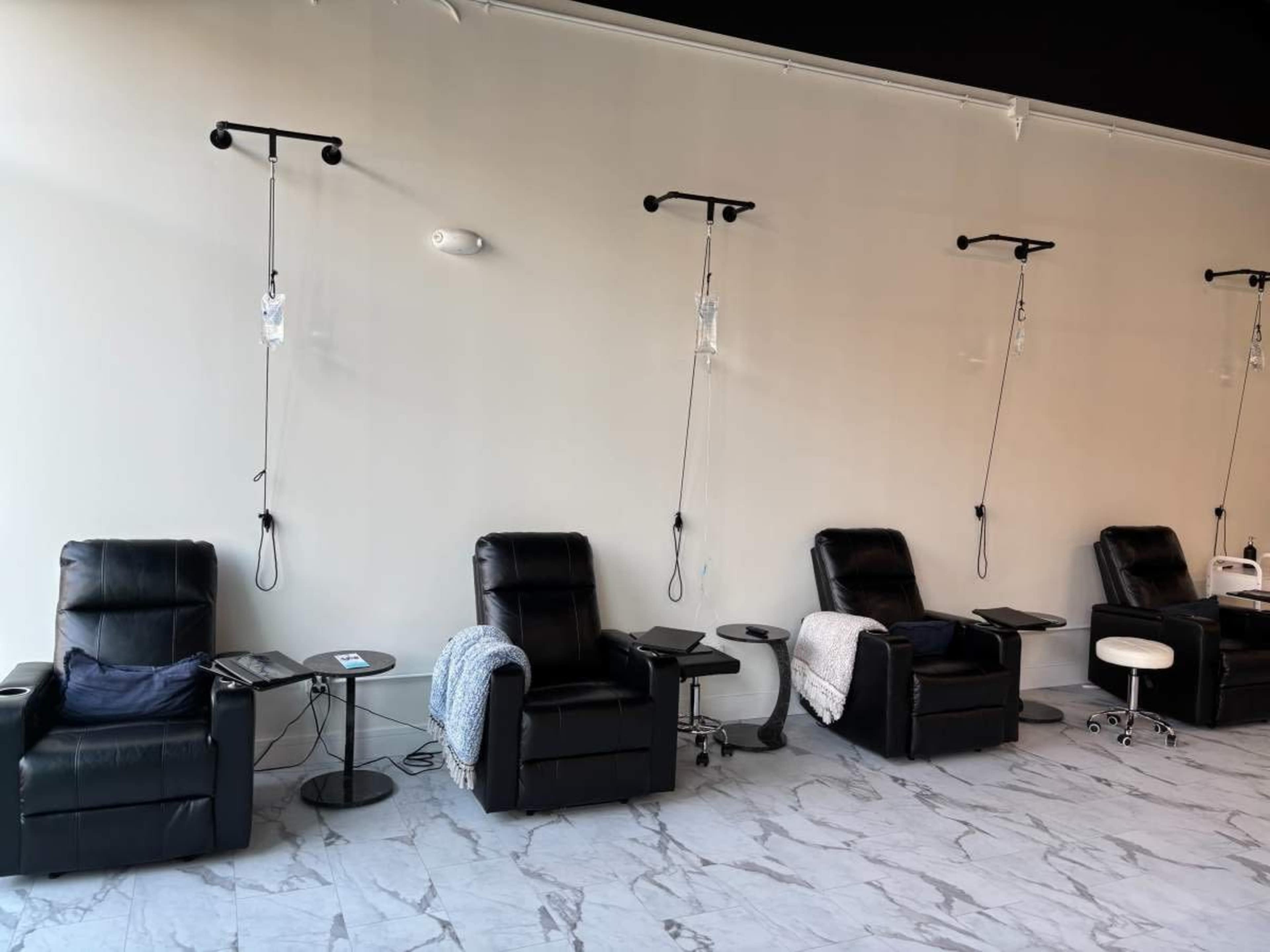 The image shows a row of black reclining chairs with IV drip setups in a minimalist room with marble flooring.
