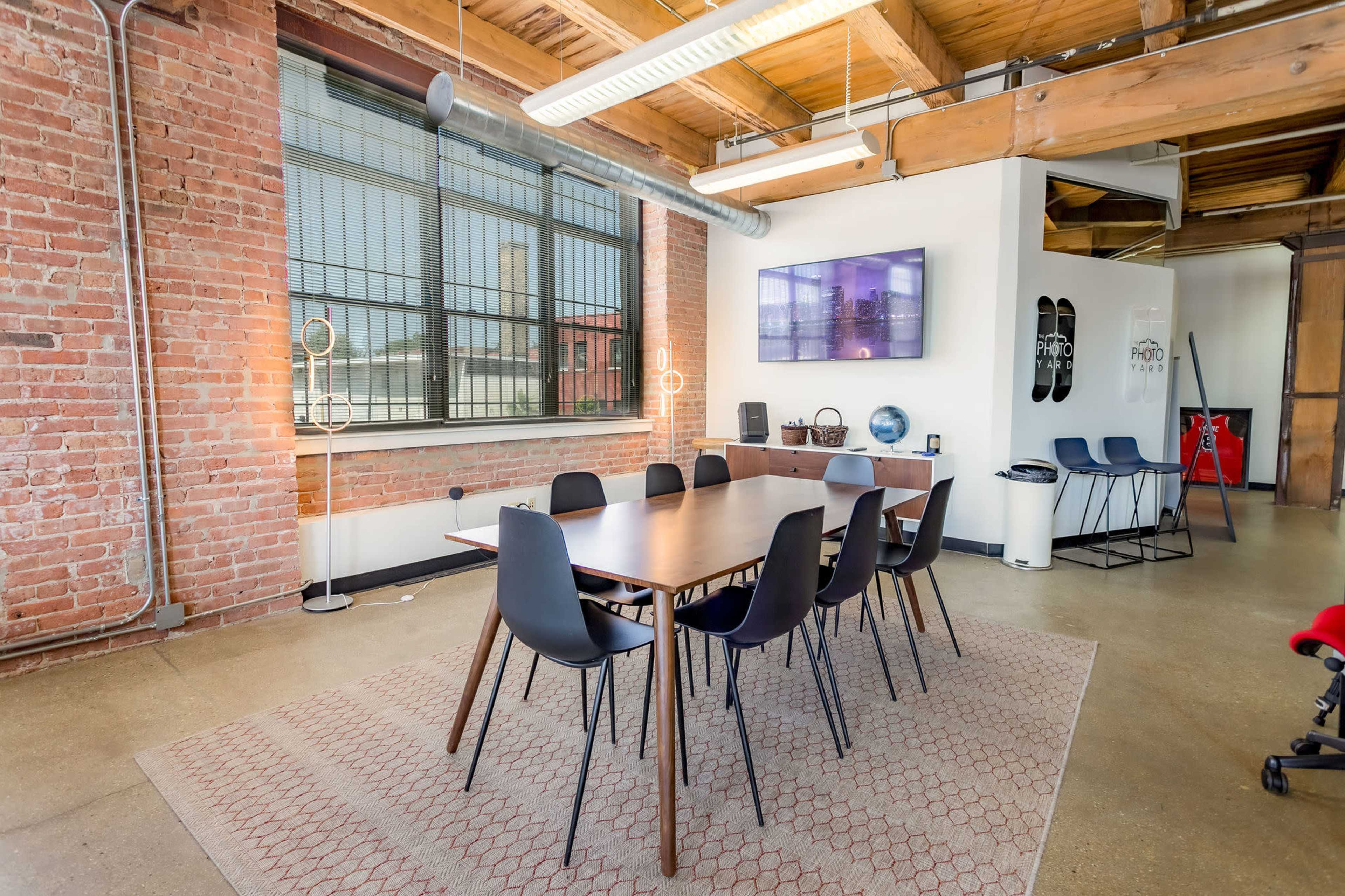 A conference room features a long wooden table surrounded by black chairs, with exposed brick walls and large windows.