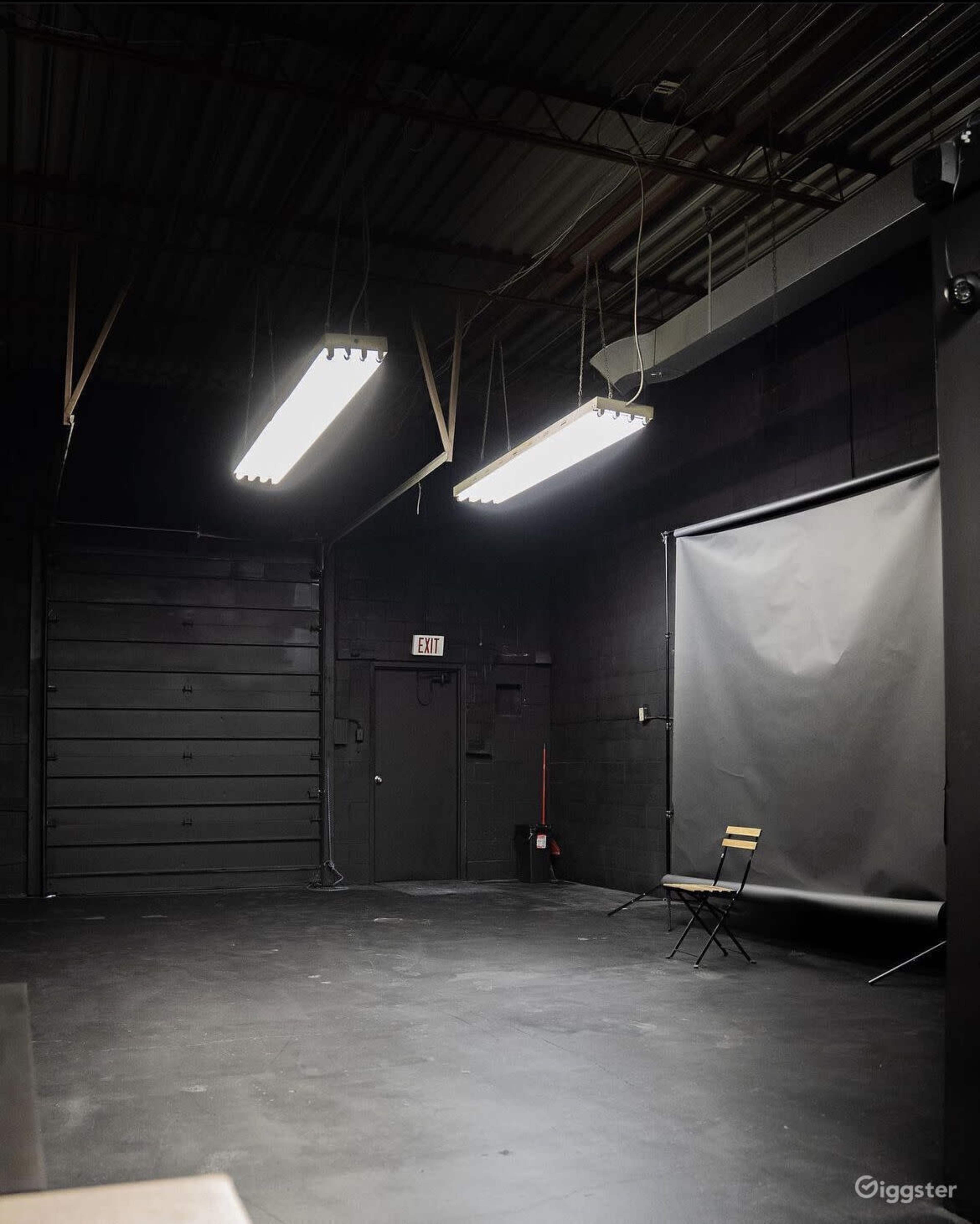 The image shows an empty, dimly lit studio space with a large gray backdrop, a single chair, and overhead fluorescent lights.