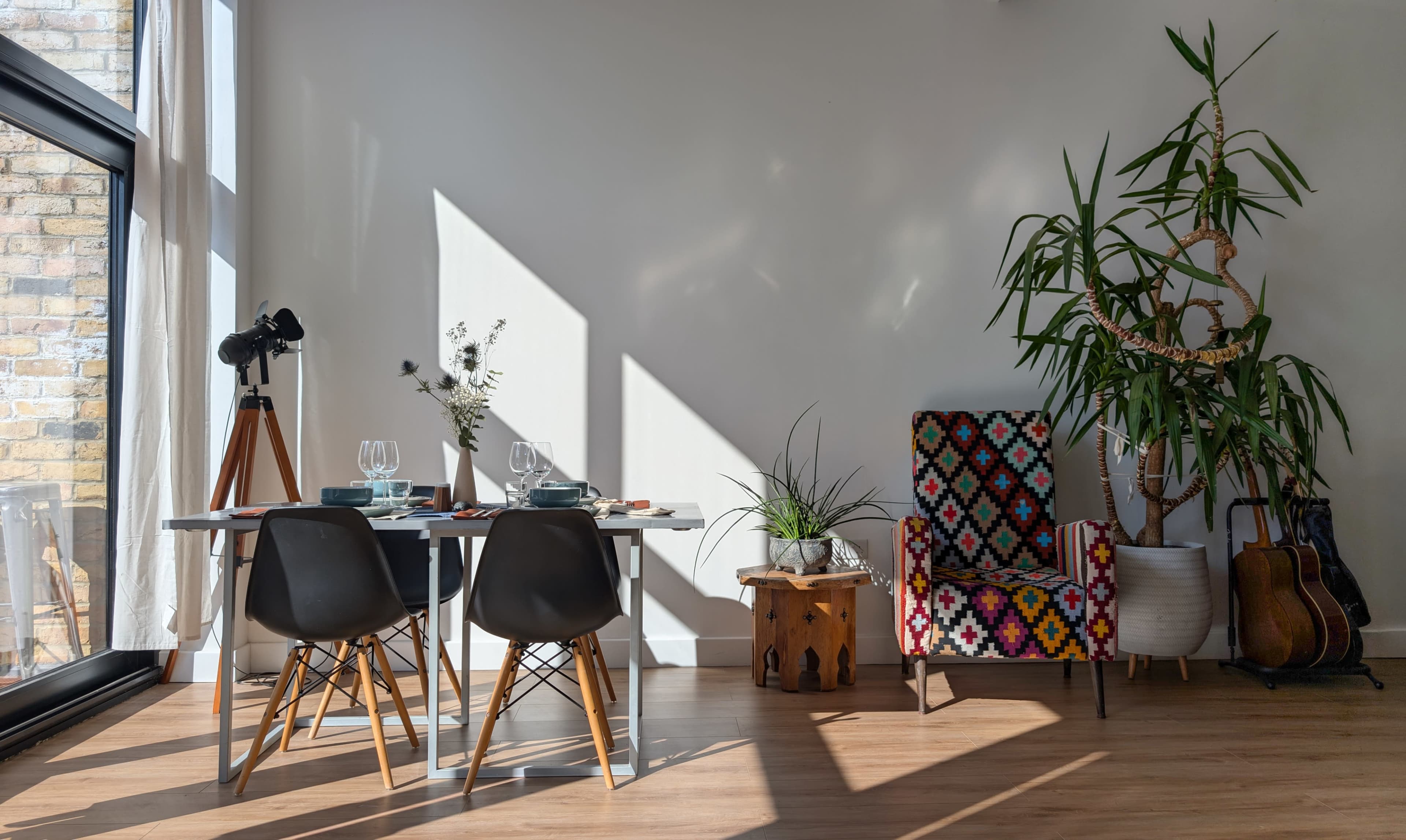 A dining area features a table set for two with a telescope nearby, a colorful chair, a potted plant, and sunlight casting shadows across the wall.
