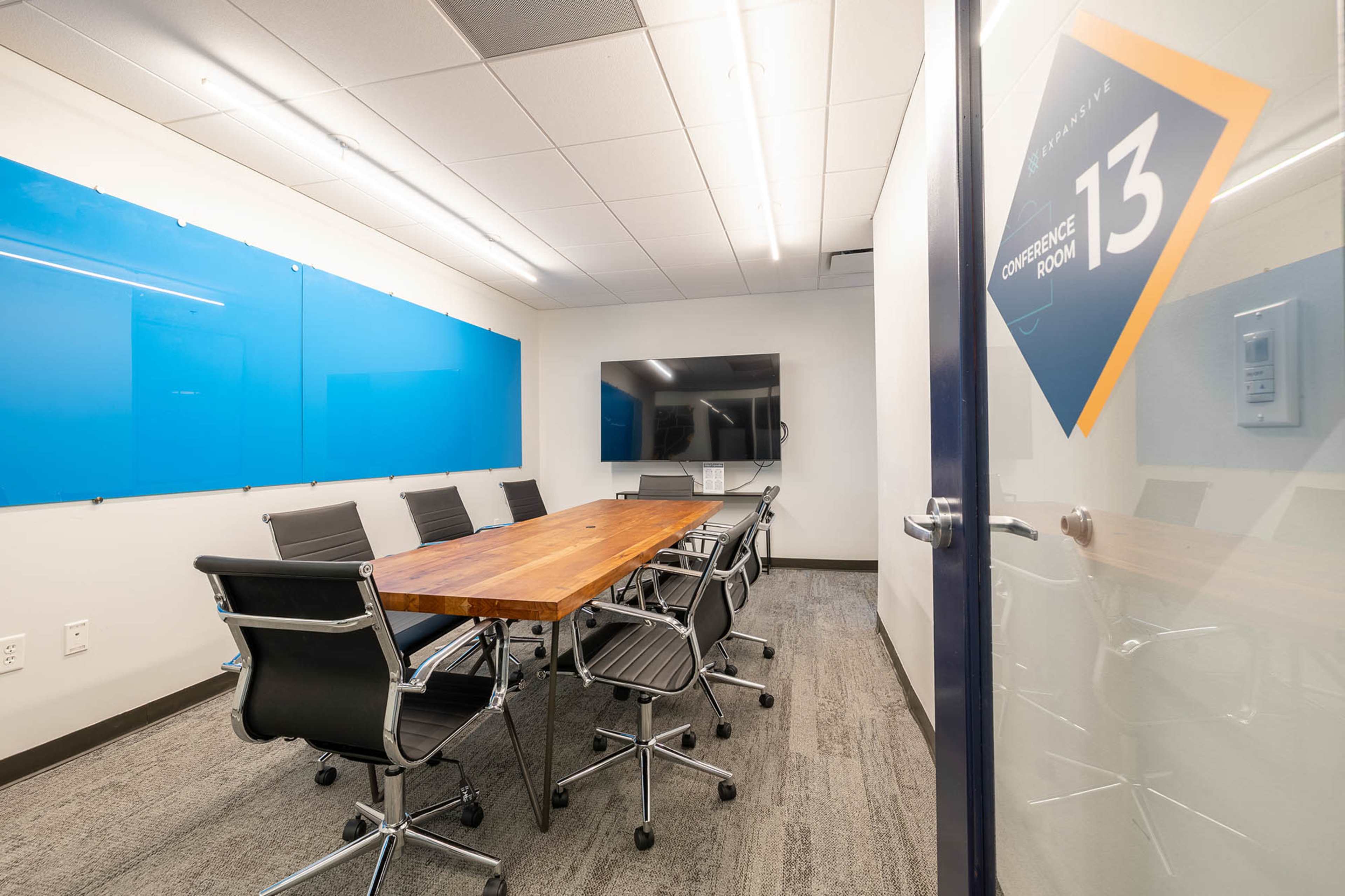 A modern conference room features a long wooden table surrounded by black chairs, a large screen on the wall, and blue presentation boards.