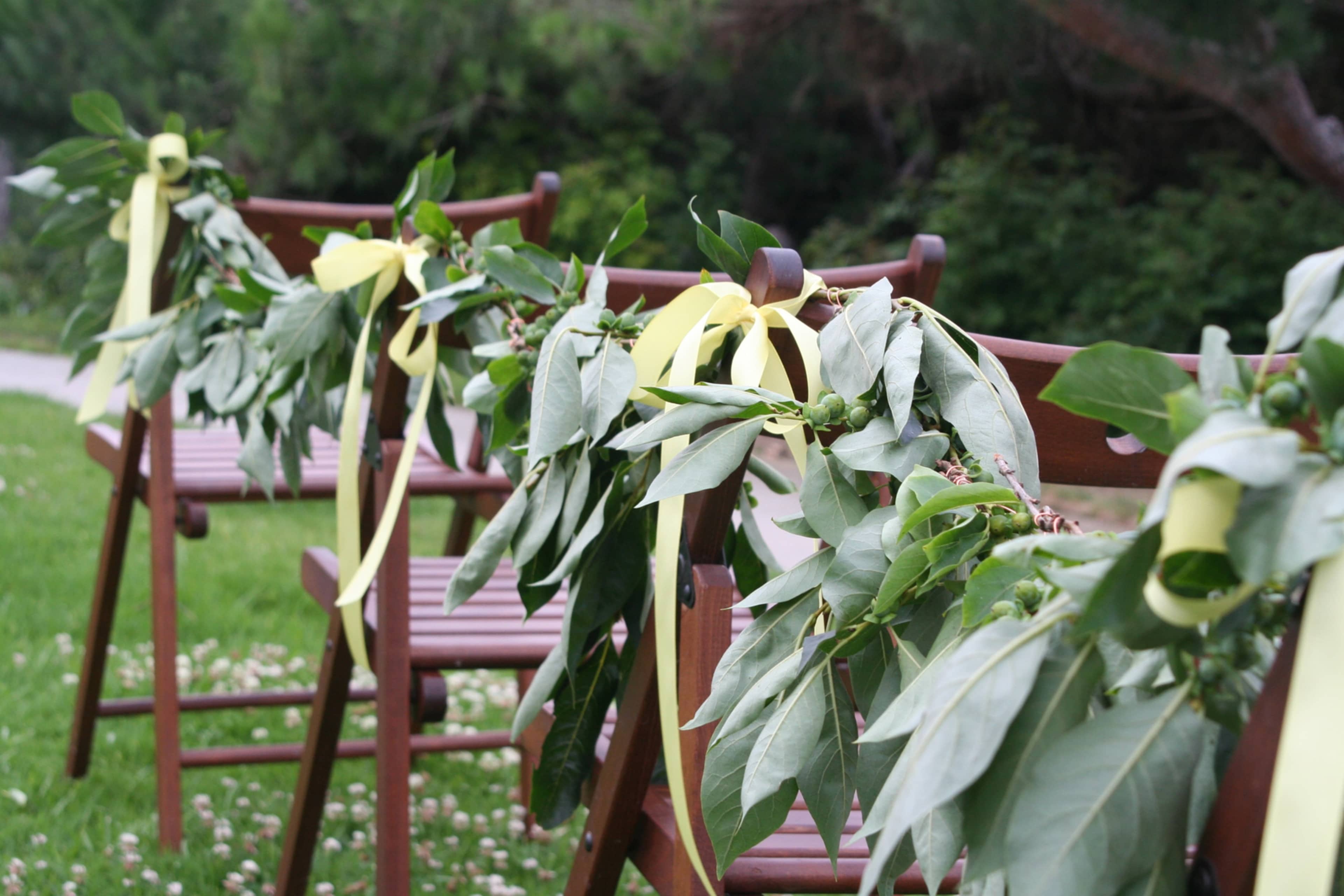 Wooden chairs are decorated with leafy garlands and yellow ribbons in a grassy outdoor setting.