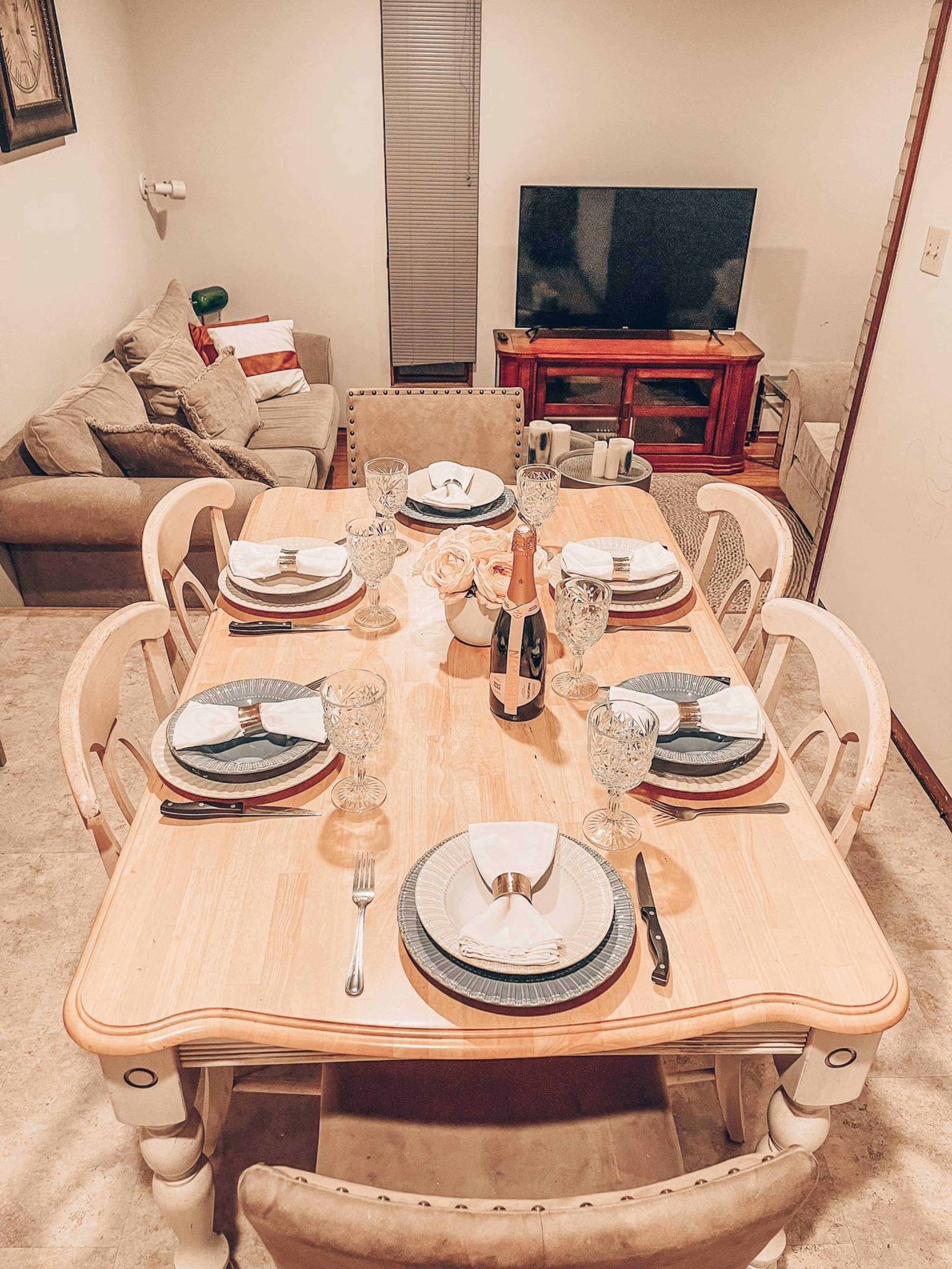 A dining table is set for a meal with plates, glasses, utensils, and a bottle of champagne, while a couch and television are visible in the background.