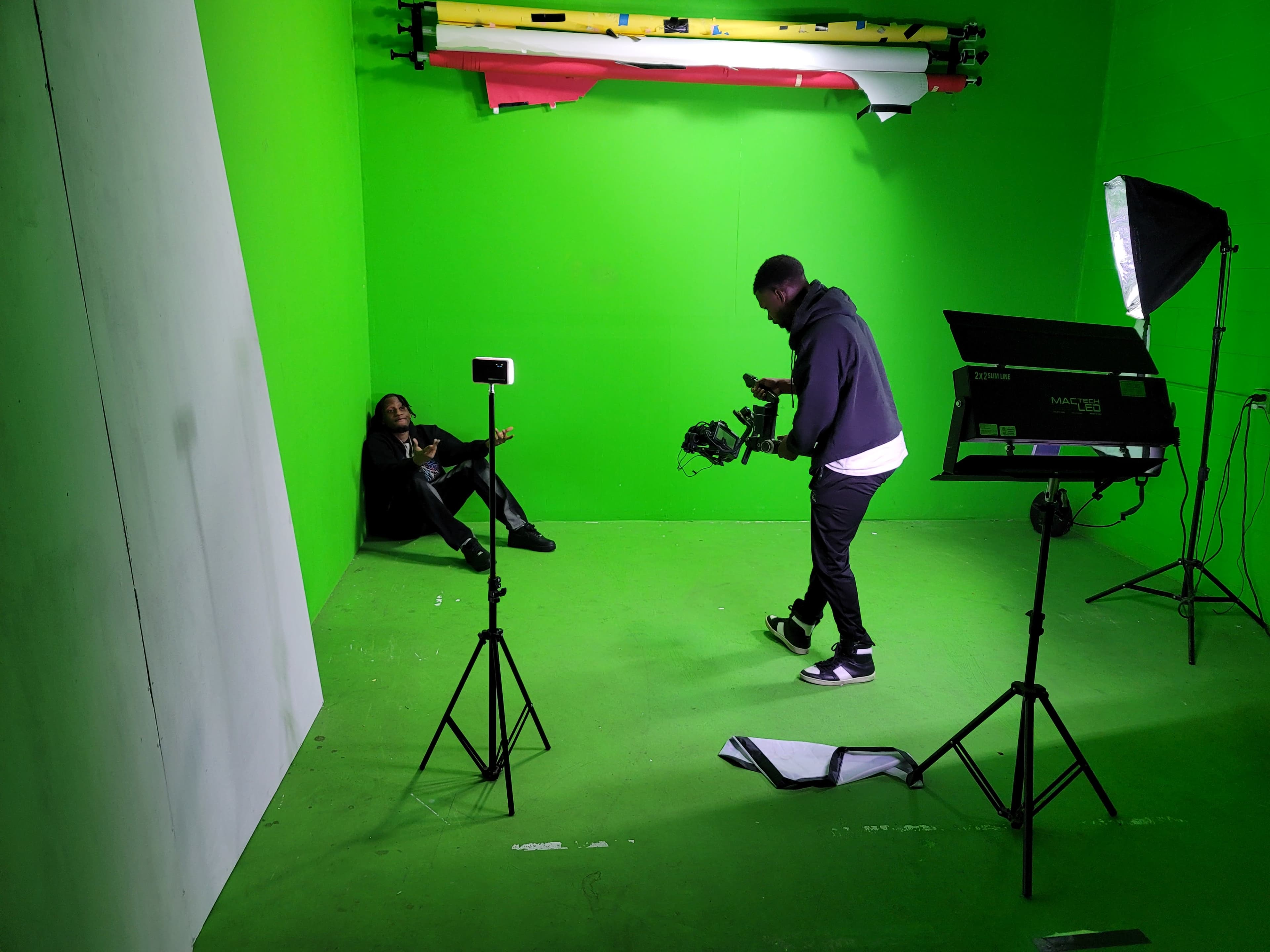 A person holding a camera stands next to an individual seated on the floor in a green screen studio setting.