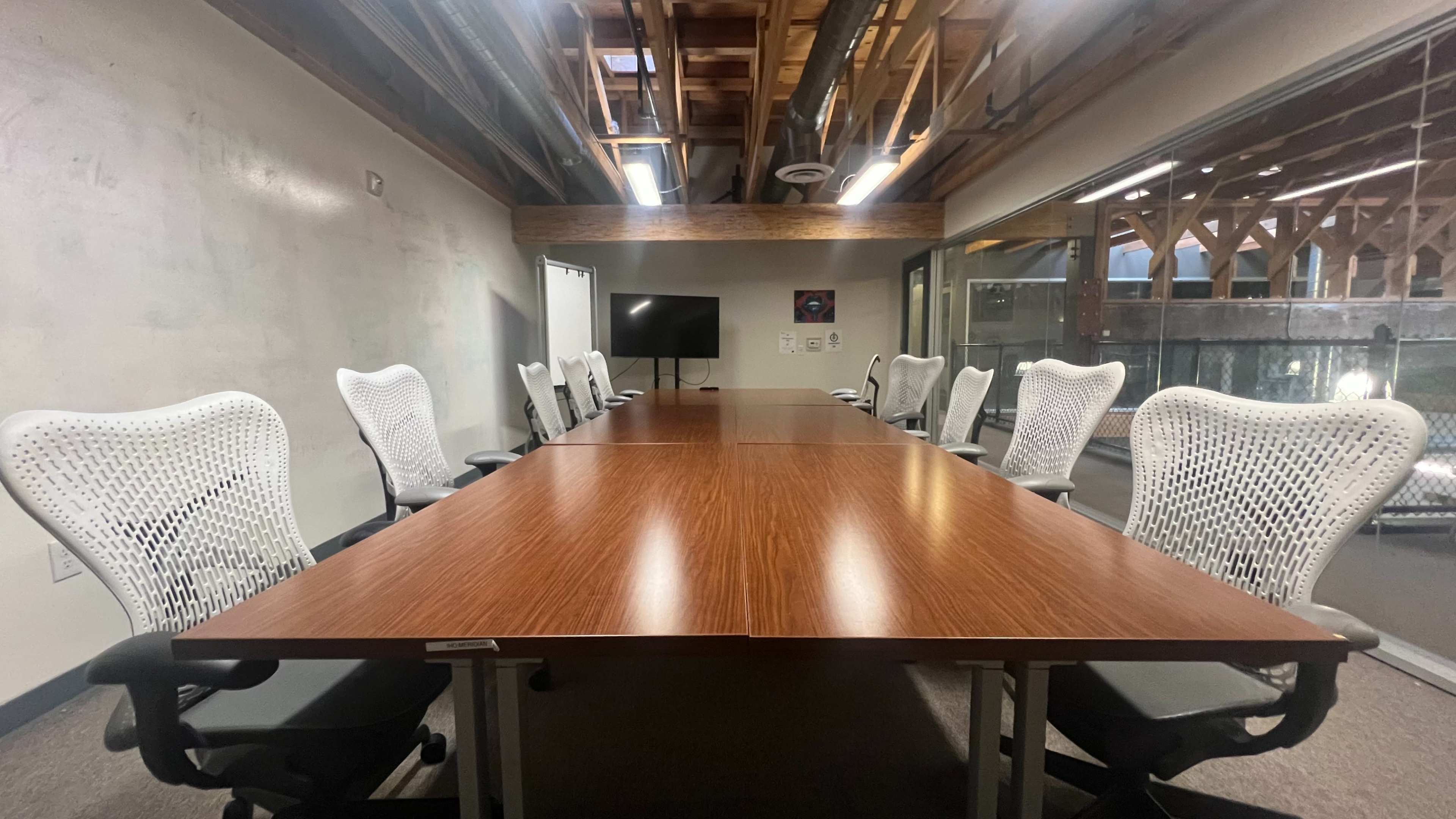 A conference room features a long wooden table surrounded by ergonomic white chairs under a ceiling with exposed beams.