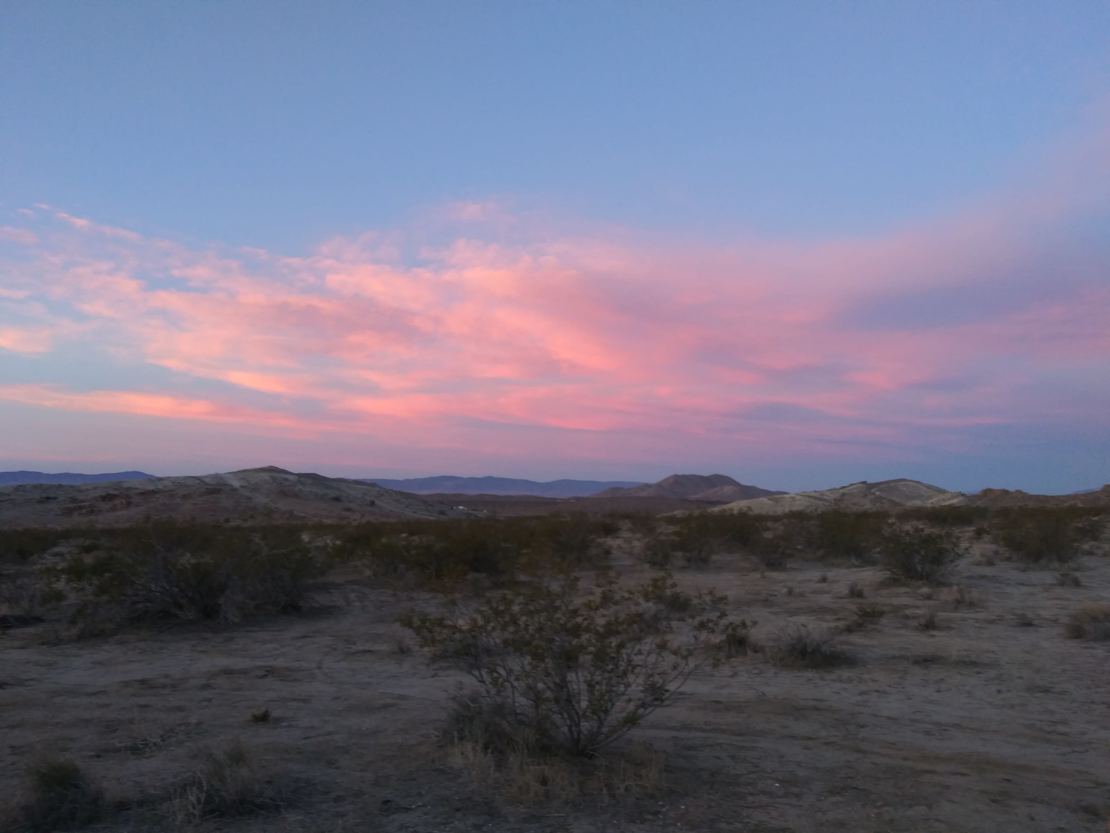 A desert landscape at dusk with pink clouds scattered across the sky above low hills and sparse vegetation.