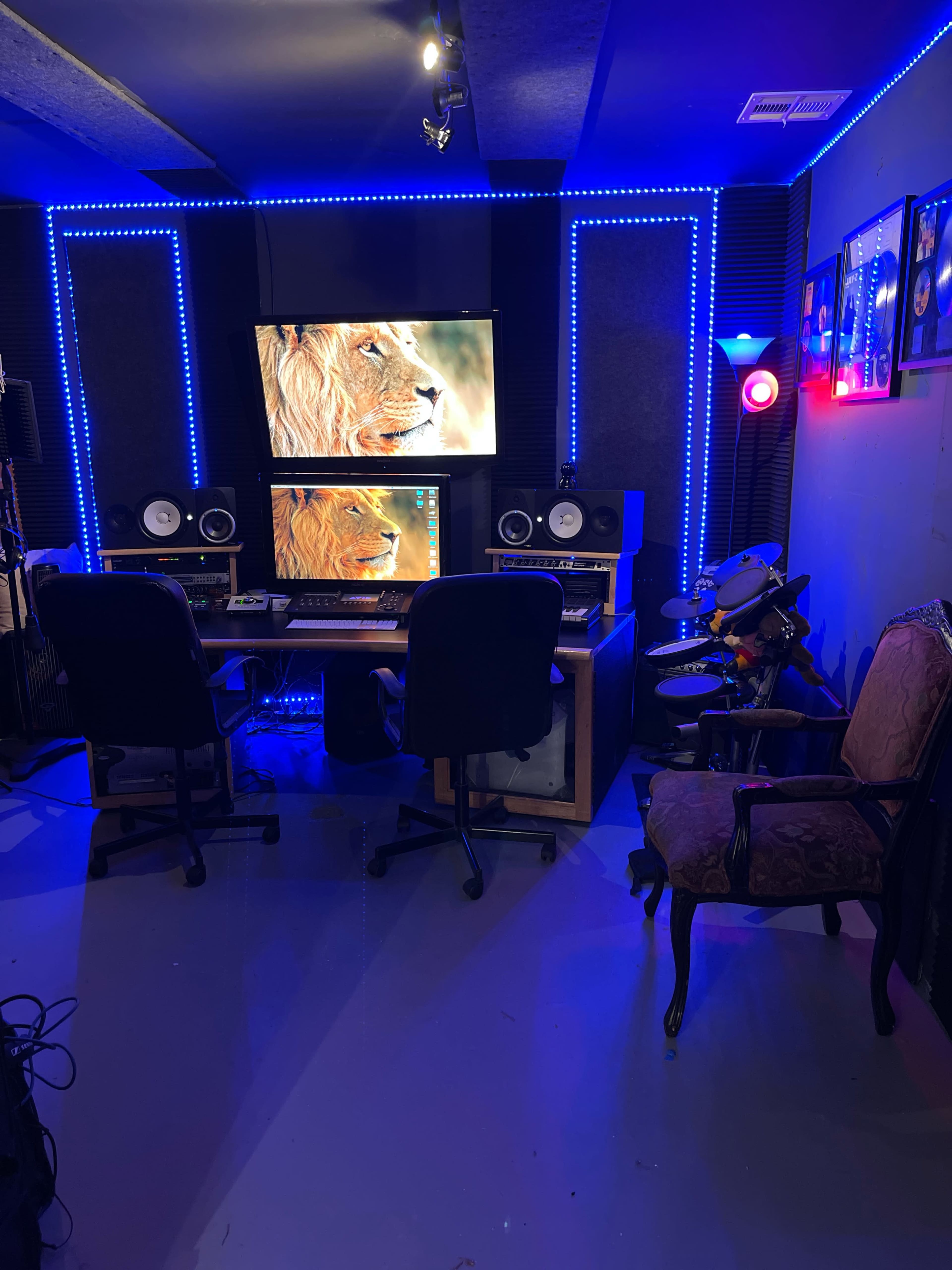 A music studio with a computer desk, two chairs, and equipment surrounded by blue LED lights and decorated walls.